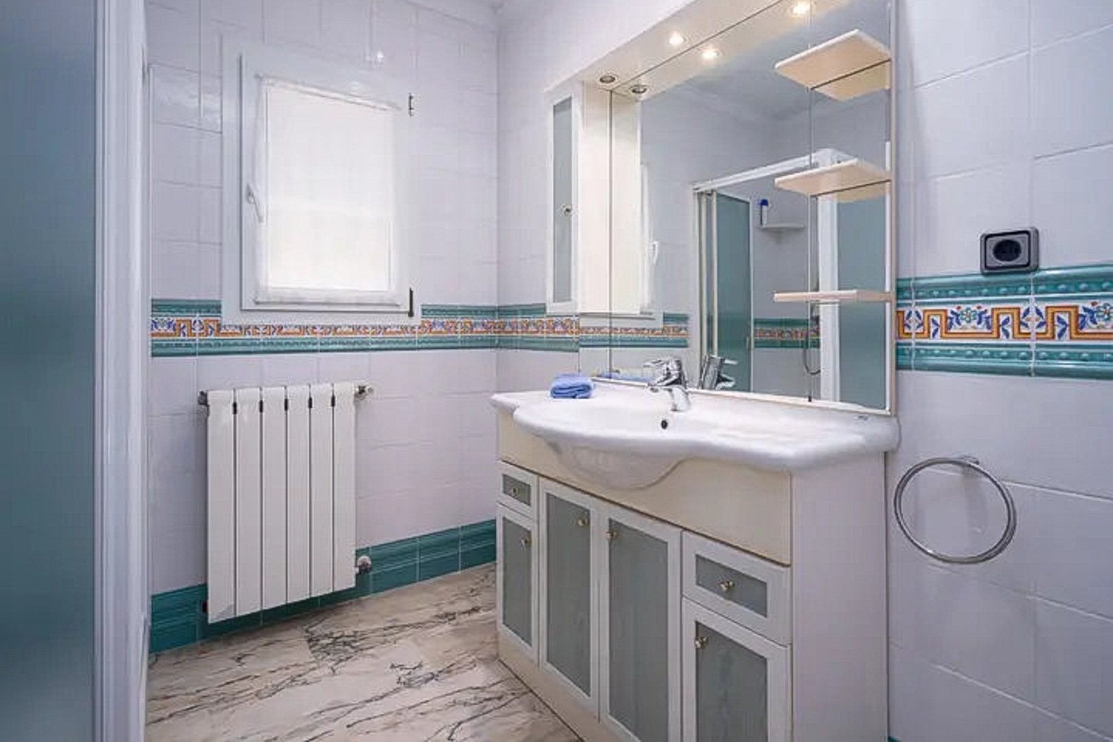 Baño con azulejos blancos y cenefa decorativa mediterránea, mueble de lavabo con puertas de cristal esmerilado, espejo con iluminación integrada y suelo de mármol veteado.