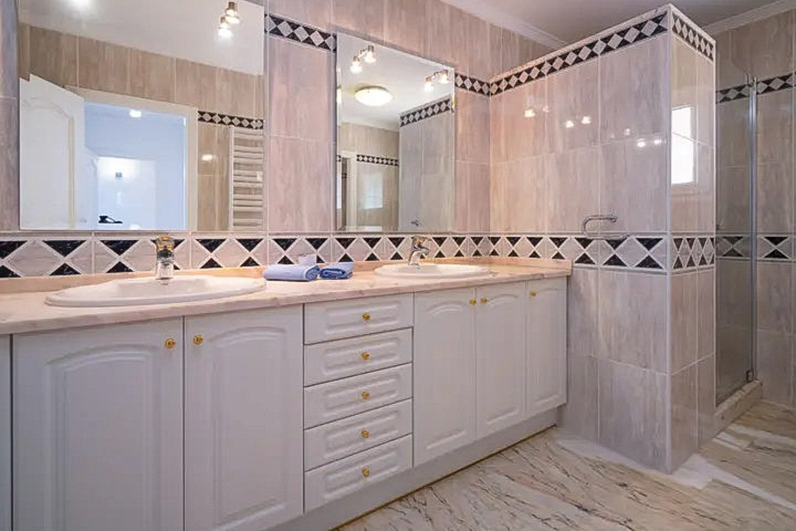 Baño con lavabo doble, mueble blanco de estilo clásico y encimera de mármol. Destacan los azulejos con cenefa de rombos y la zona de ducha integrada.