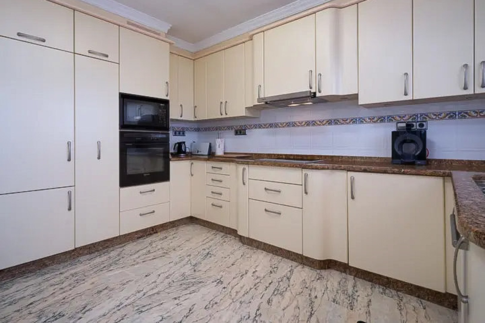 Cocina equipada con armarios en tono crema, encimera de granito marrón y electrodomésticos integrados. Destaca el suelo de mármol veteado y el detalle de azulejos con cenefa mediterránea.