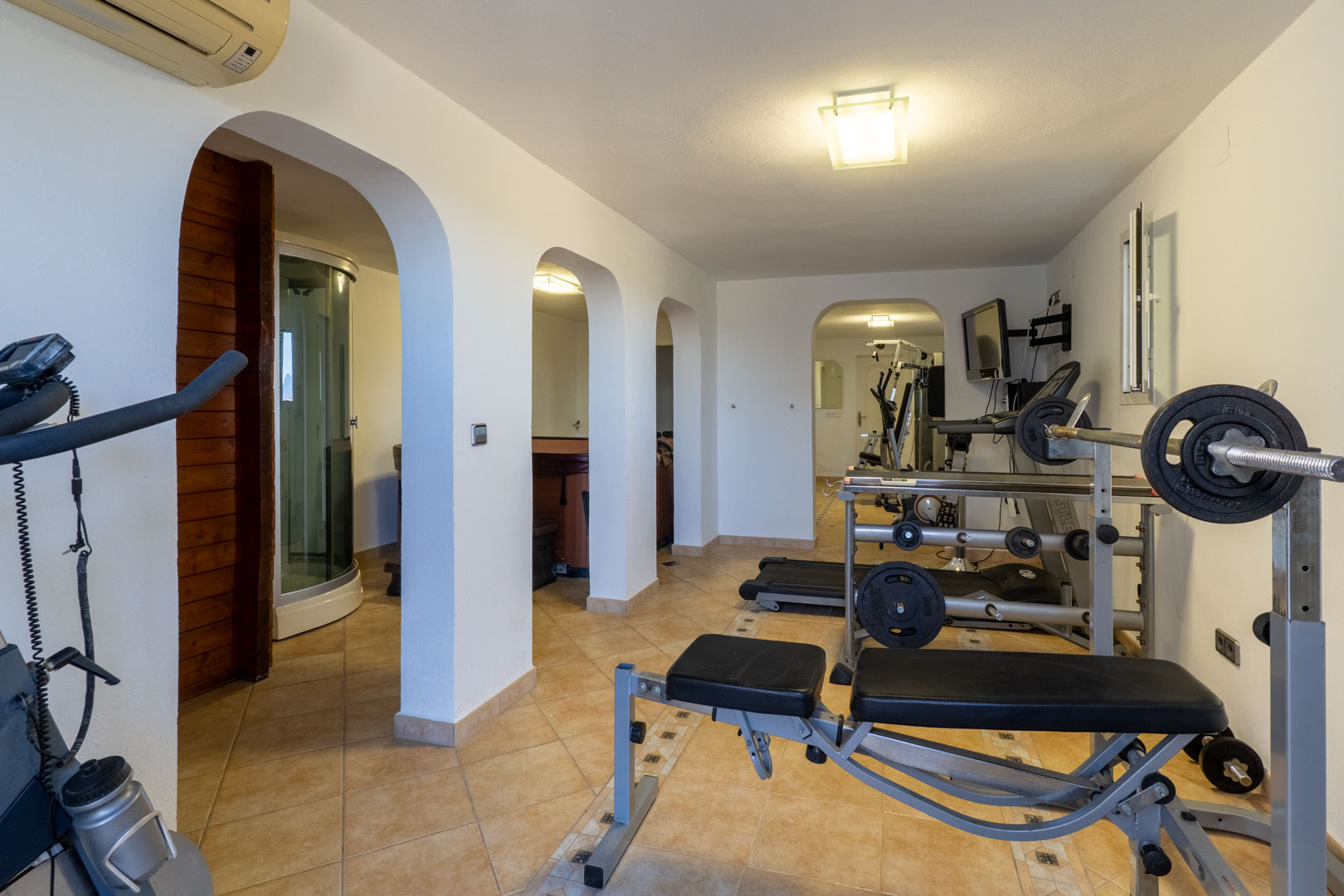 Gimnasio privado y zona de bienestar con máquinas de musculación, cinta de correr y acceso a sauna y ducha de vapor mediante elegantes arcos mediterráneos.