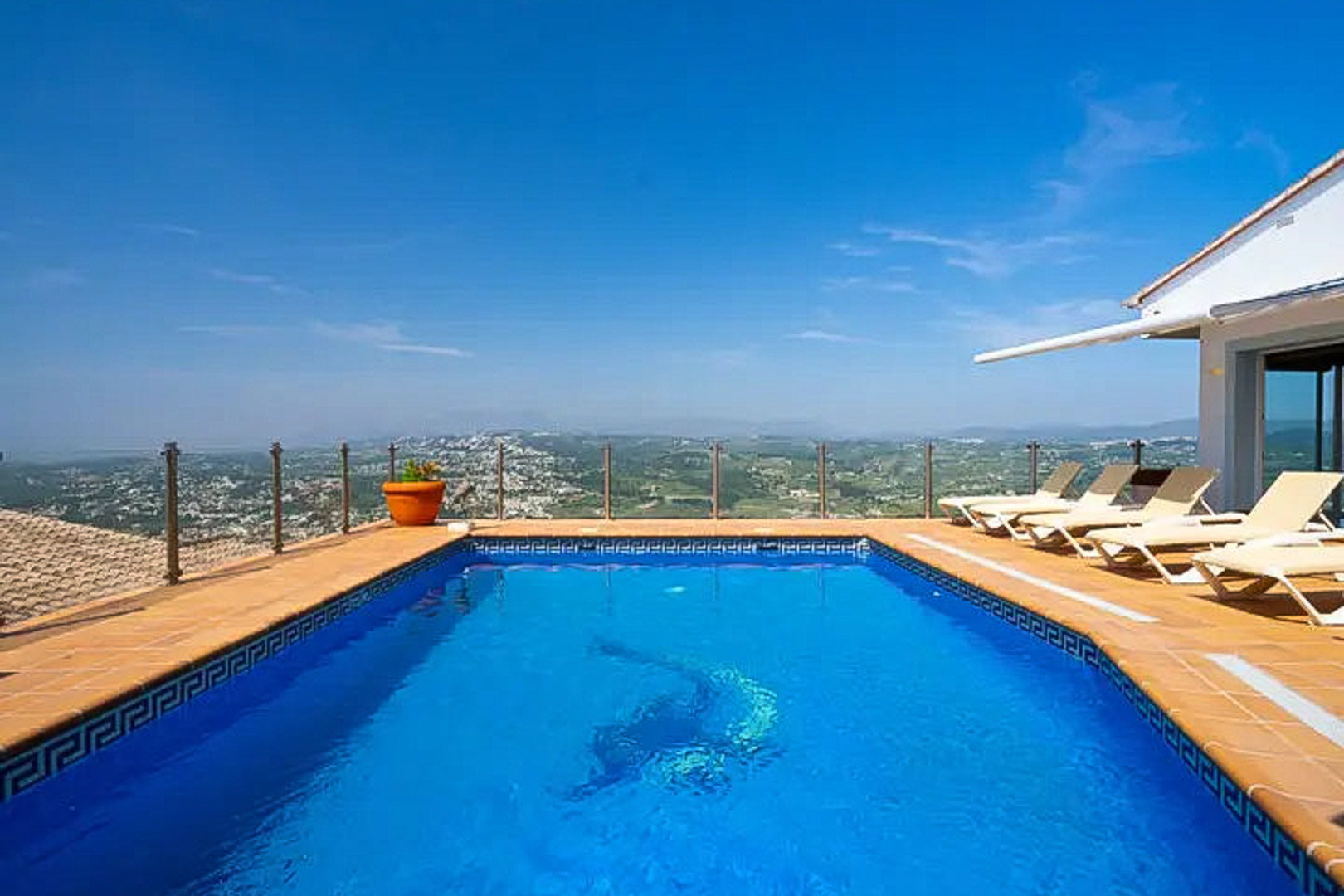 Piscina rectangular con mosaico de delfín, rodeada de una terraza con tumbonas y barandilla de cristal que ofrece vistas panorámicas al valle y la costa.