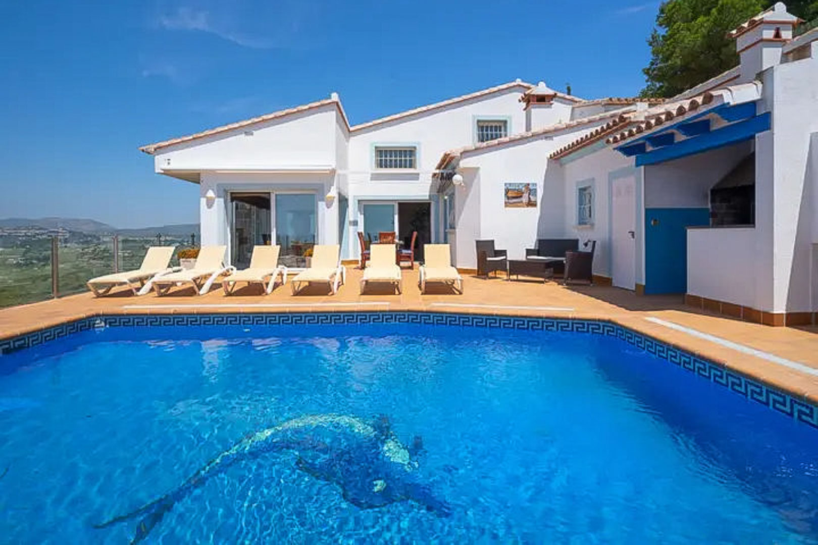 Terraza con piscina decorada con mosaico de delfín, zona de solárium con tumbonas y área de barbacoa bajo vigas azules, con vistas despejadas al paisaje.