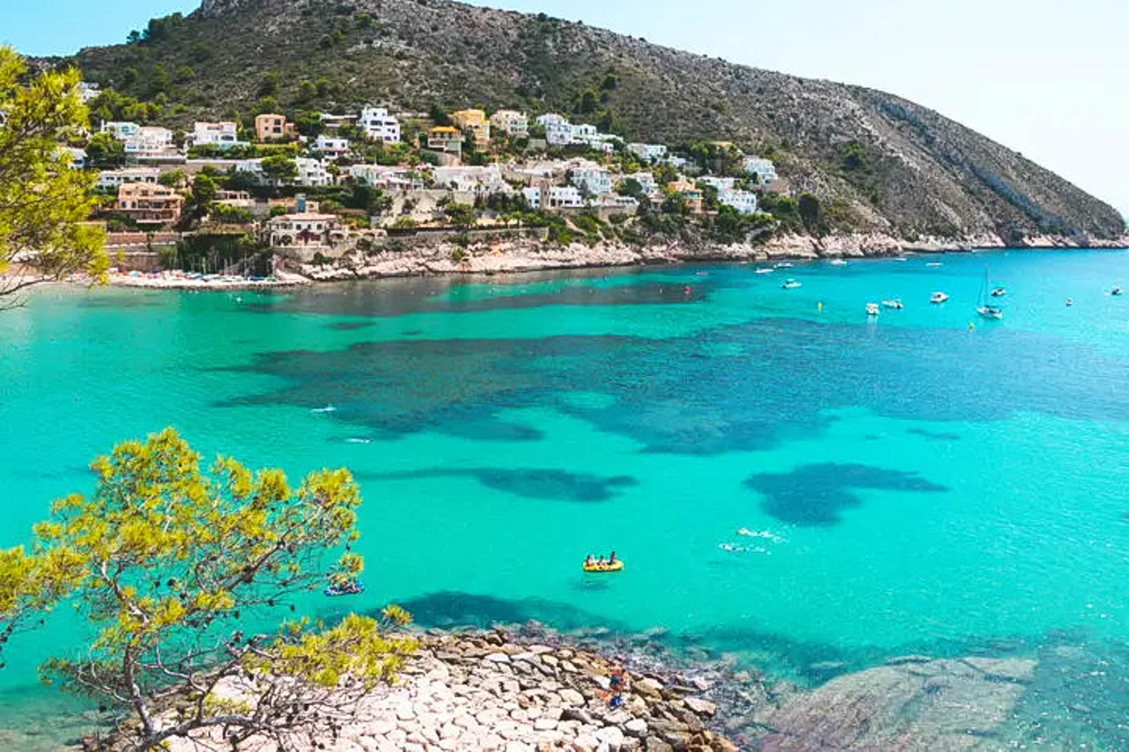 Vista panorámica de una cala en Moraira con aguas cristalinas color turquesa, rodeada de pinos y villas blancas integradas en la ladera de la montaña.