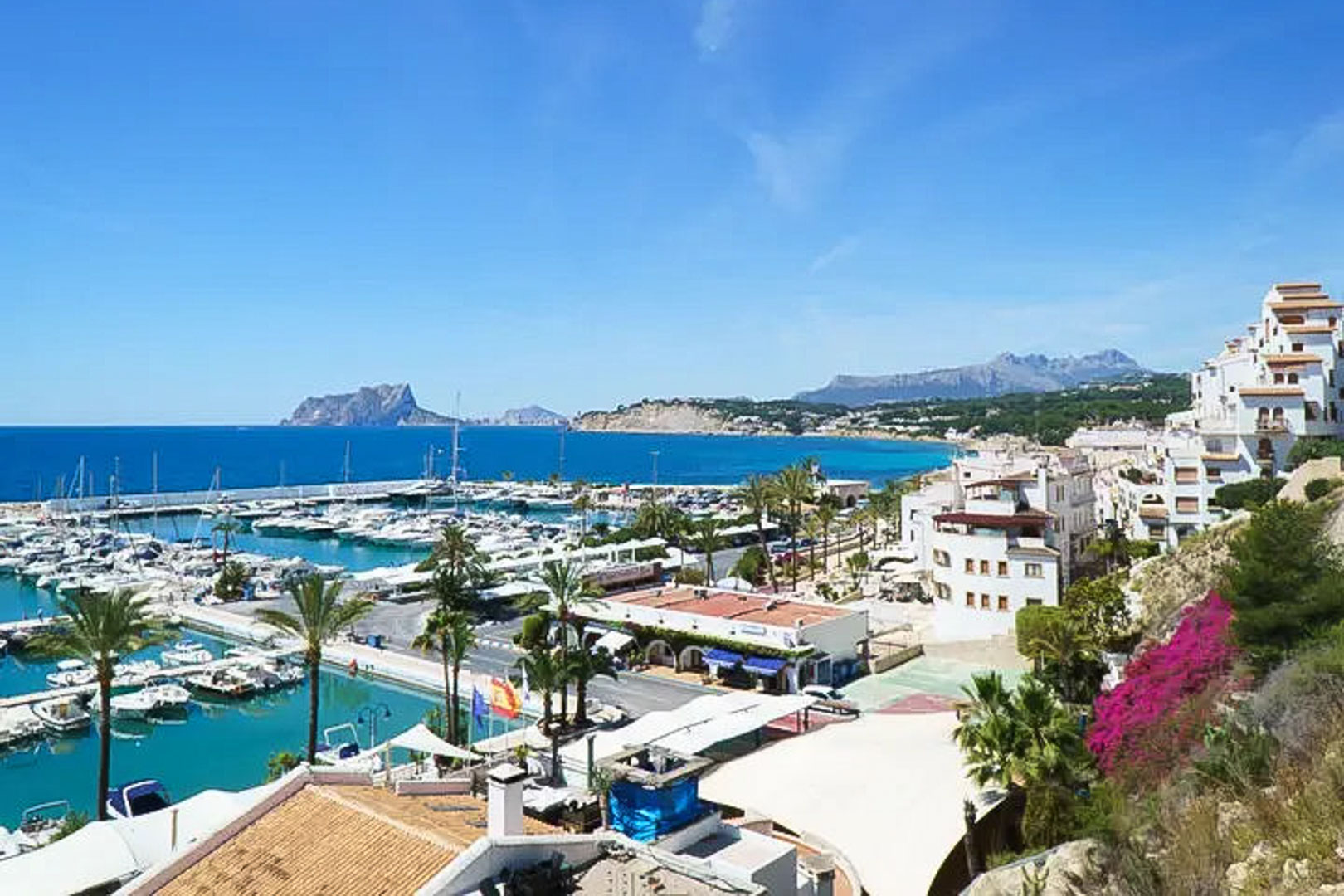 Vista panorámica del puerto deportivo de Moraira con el Peñón de Ifach al fondo, rodeado de arquitectura blanca tradicional y aguas turquesas del Mediterráneo.