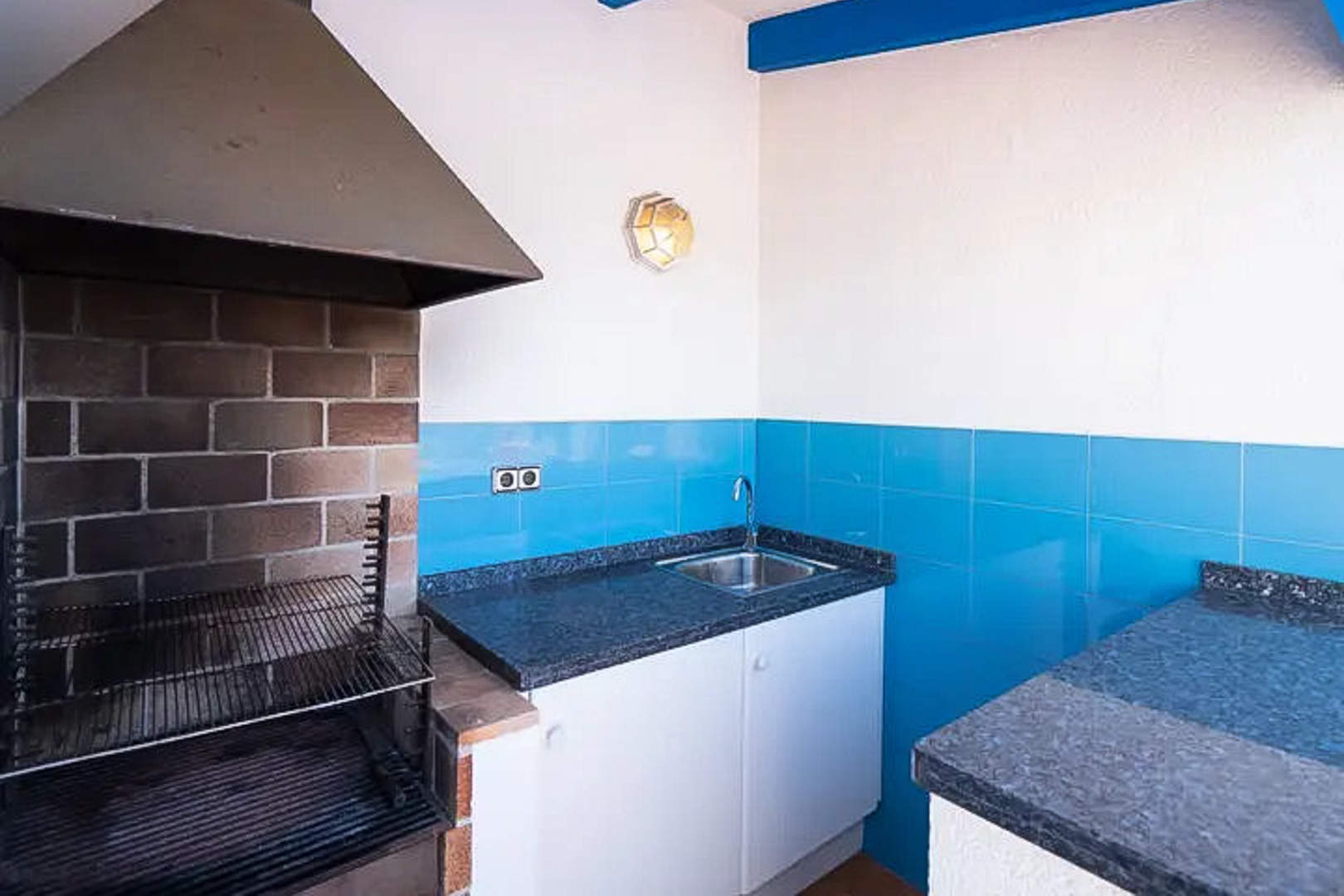 Zona de barbacoa exterior equipada con campana metálica, encimera de granito oscuro, fregadero de acero inoxidable y azulejos en azul mediterráneo.