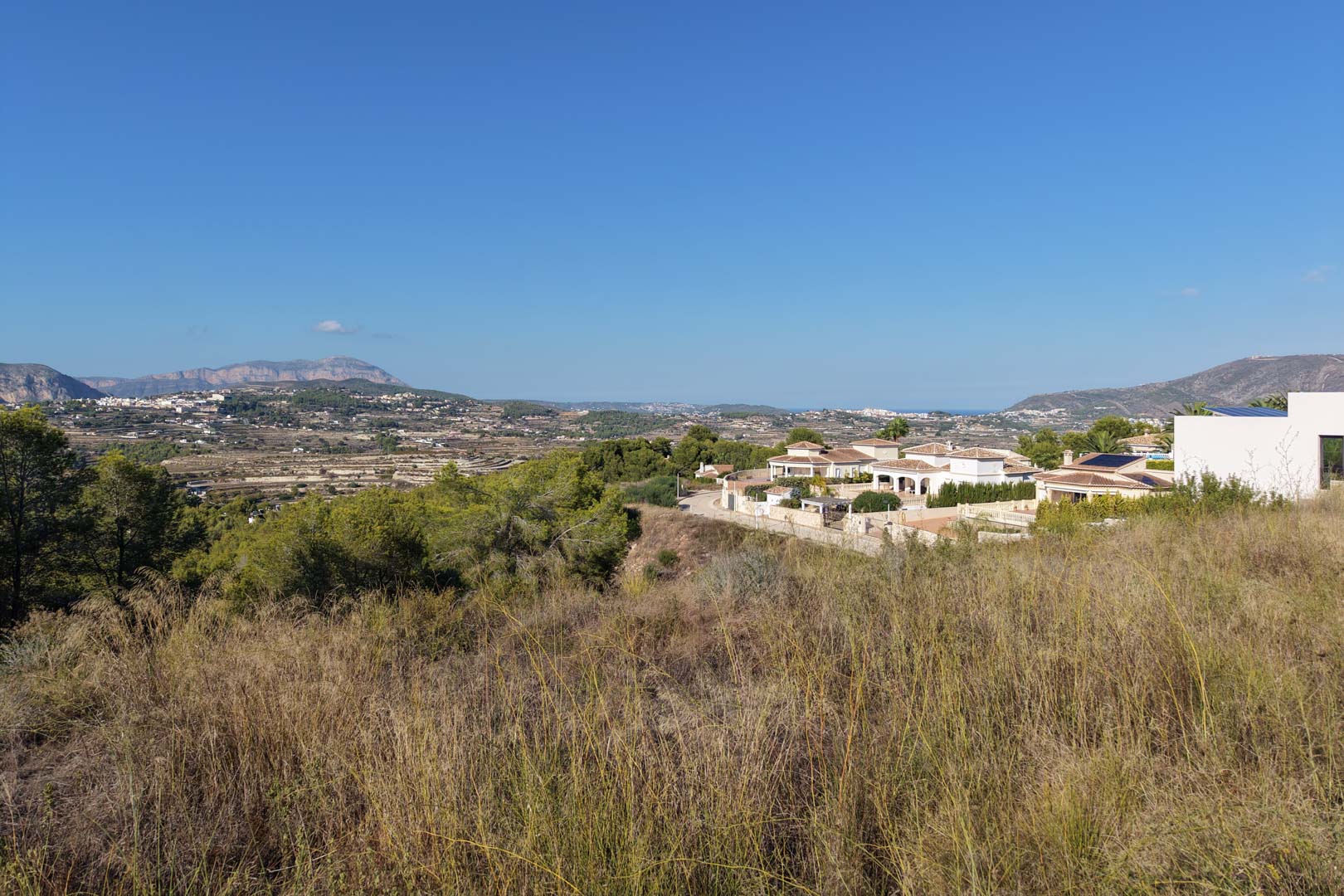 Vista elevada de la parcela urbana con vegetación silvestre, ofreciendo vistas abiertas al valle, montañas distantes y villas de estilo mediterráneo.