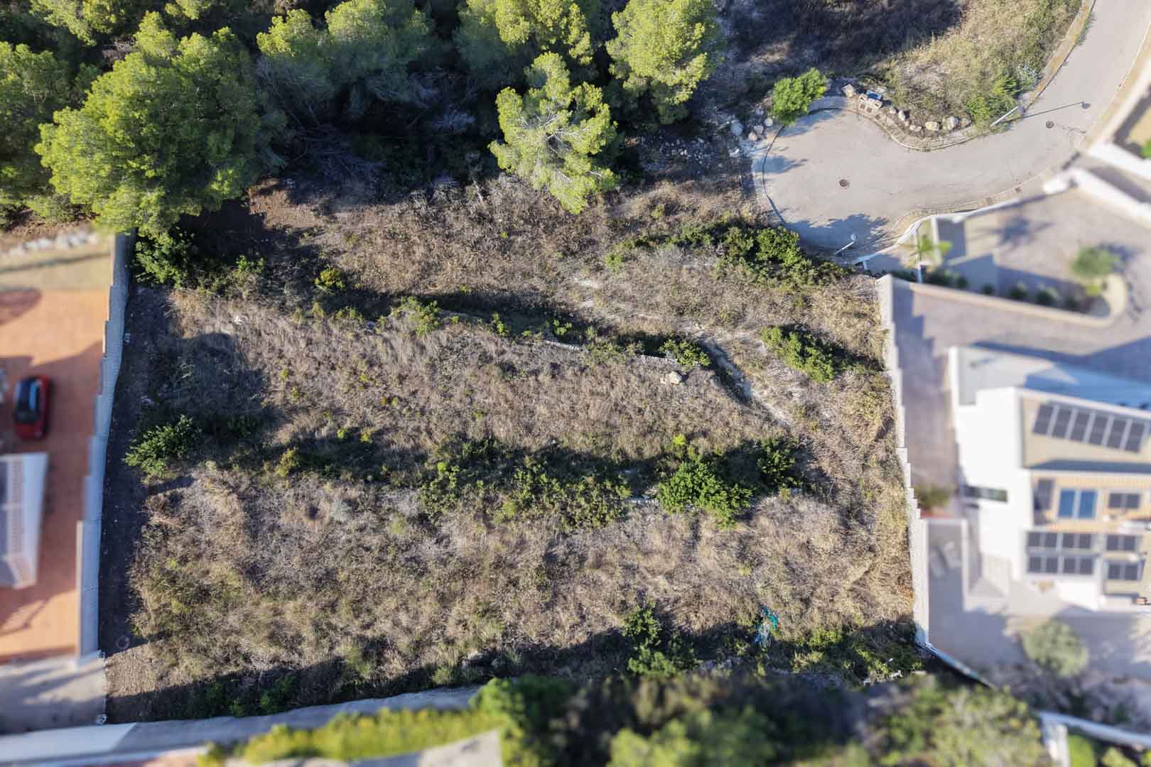 Vista aérea cenital de la parcela urbana escalonada con muros de piedra seca y vegetación mediterránea, situada junto a una zona residencial consolidada.