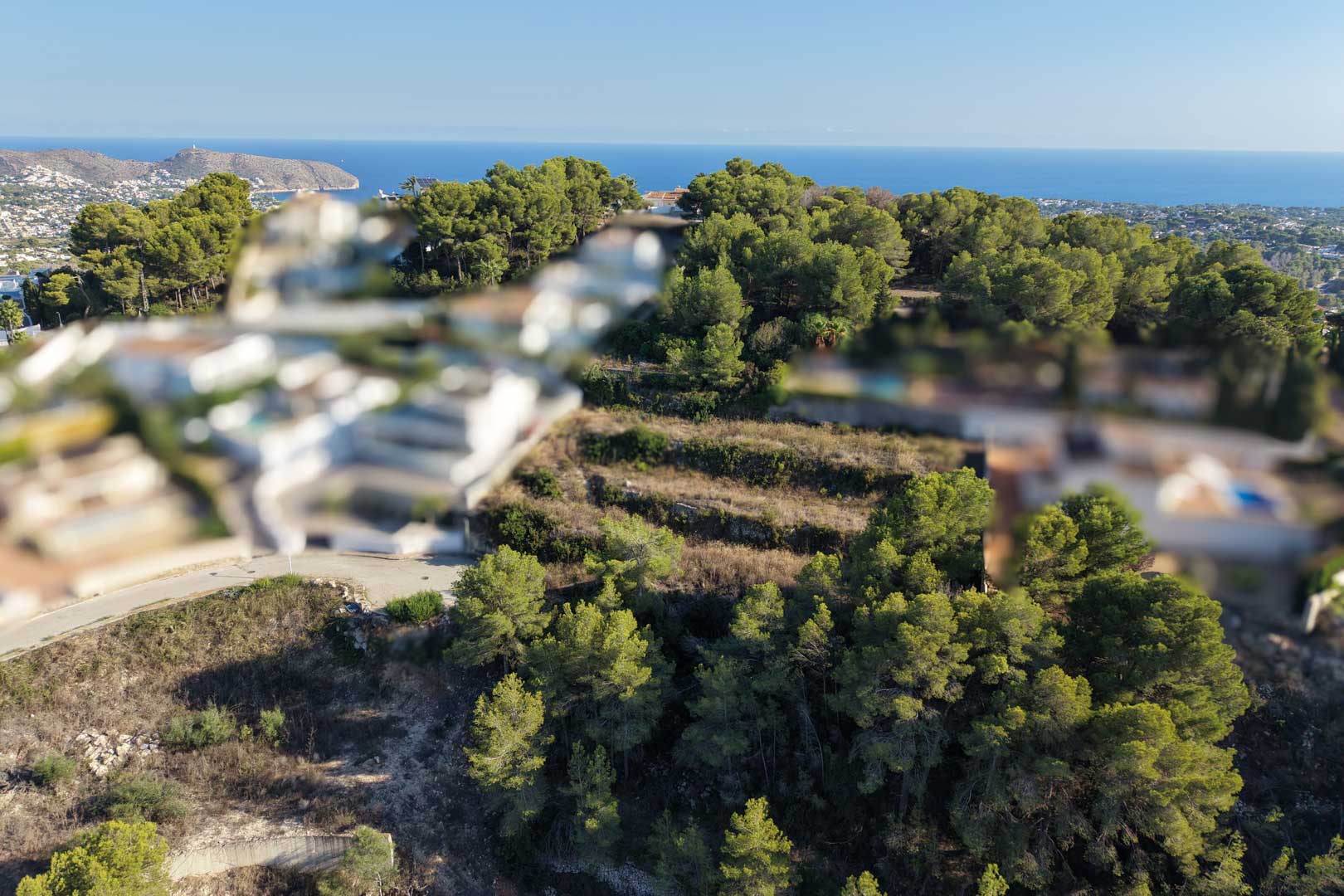 Vista aérea de una parcela urbana aterrazada en Moraira, rodeada de pinos y con vistas panorámicas al mar Mediterráneo bajo un cielo despejado.