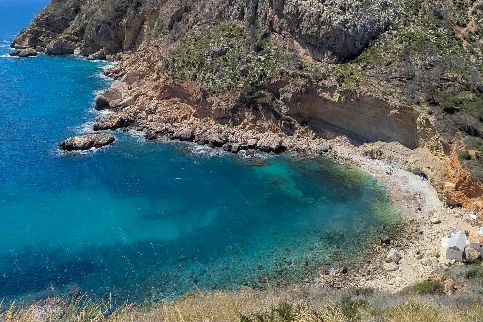 Vista aérea de una cala mediterránea con aguas turquesas, acantilados escarpados y una pequeña playa de guijarros, situada en el entorno costero de Moraira.