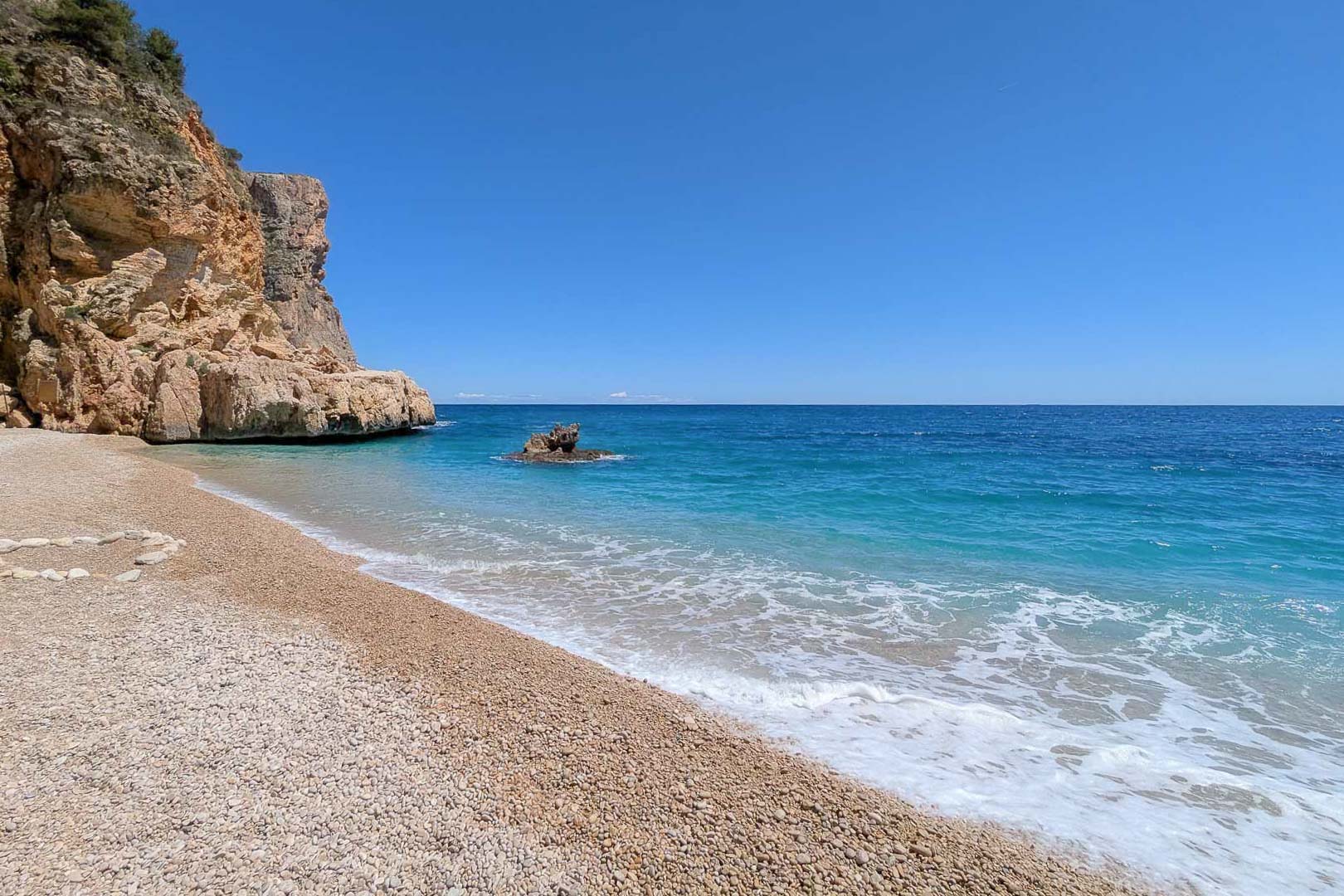 Vista de una cala de grava blanca con aguas cristalinas color turquesa, flanqueada por acantilados escarpados bajo un cielo despejado en la costa de Moraira.
