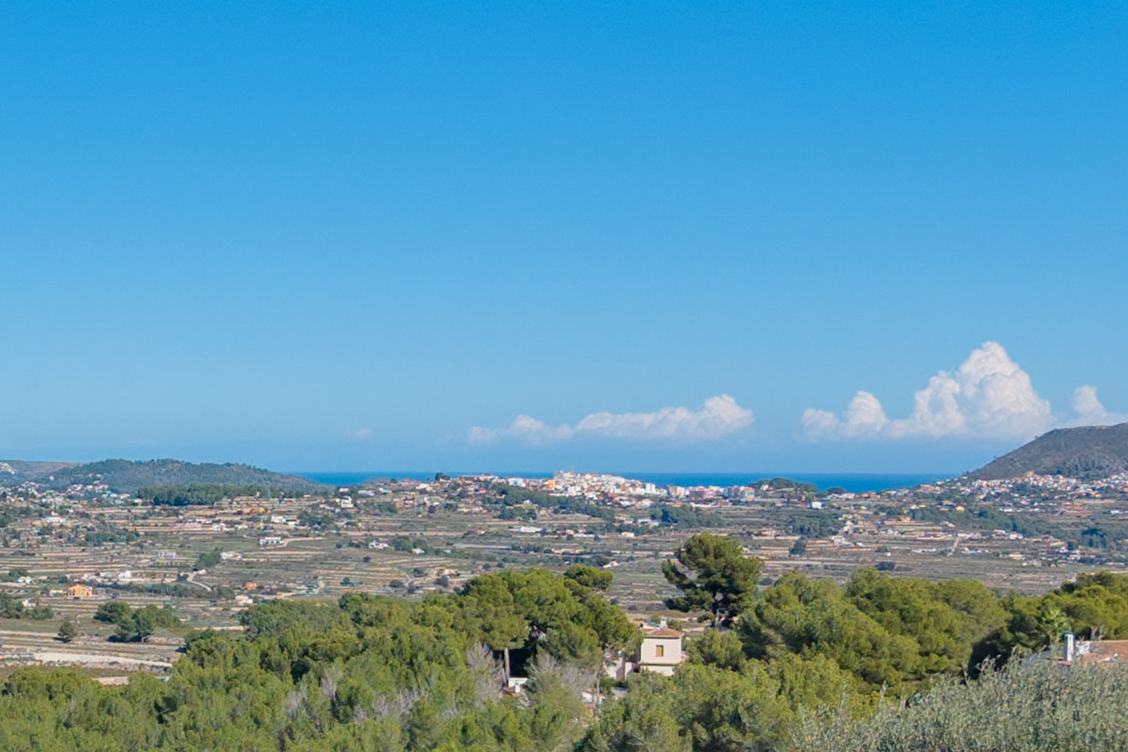 Vista panorámica desde la parcela en Benimeit, mostrando el valle aterrazado, vegetación mediterránea y una franja del mar bajo un cielo despejado.