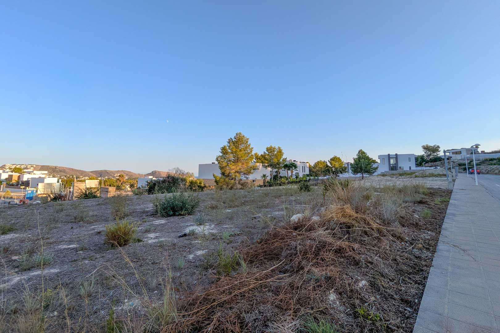 Parcela urbana llana en El Tesoro, Moraira, con vistas abiertas al valle y rodeada de villas modernas. Acceso directo desde acera pavimentada bajo un cielo despejado.