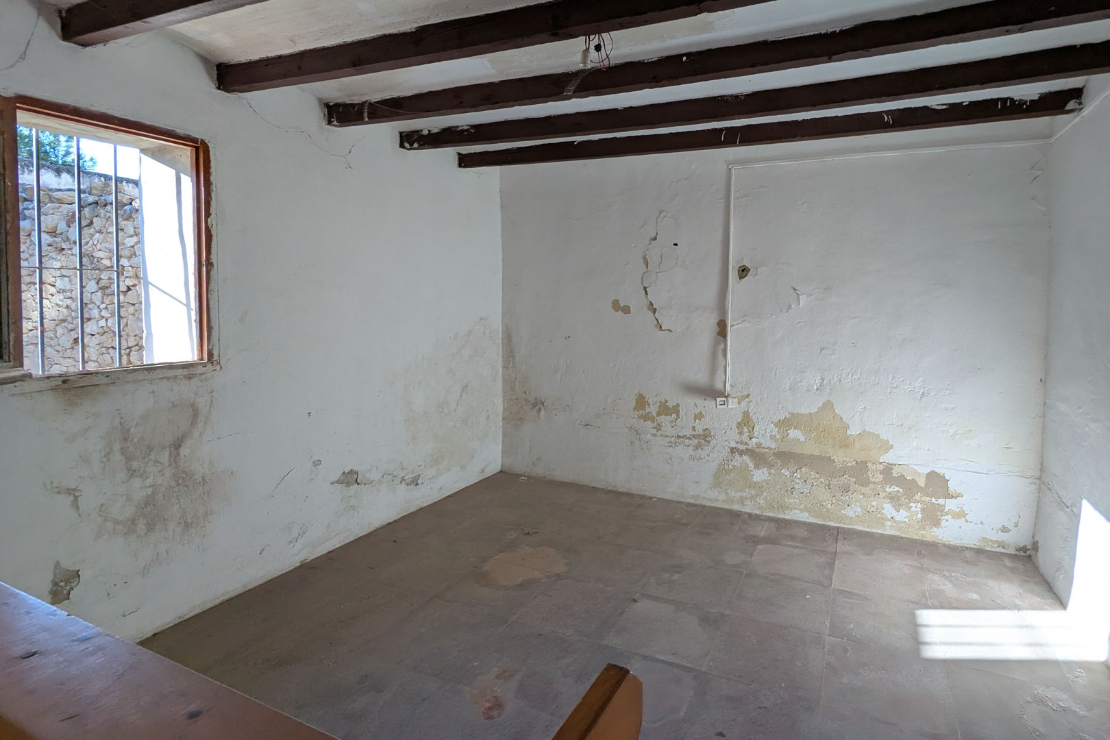 Habitación rústica con vigas de madera oscura a la vista, paredes de cal con pátina original y suelo de baldosas. La ventana con reja ofrece luz natural y vistas a un muro de piedra exterior.