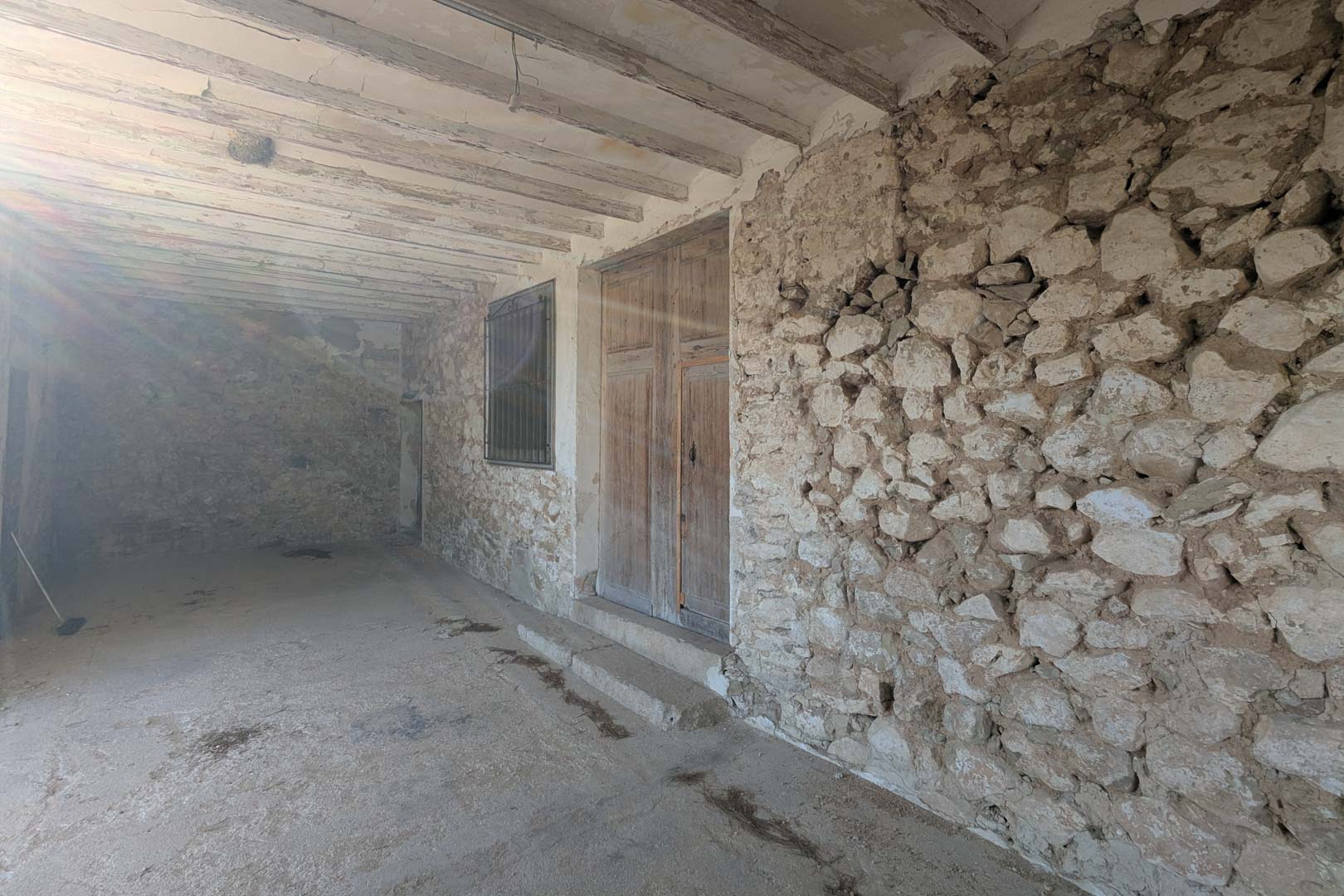 Porche cubierto con muros de piedra natural vista, vigas de madera originales y una gran puerta doble de madera antigua que da acceso a la vivienda tradicional.