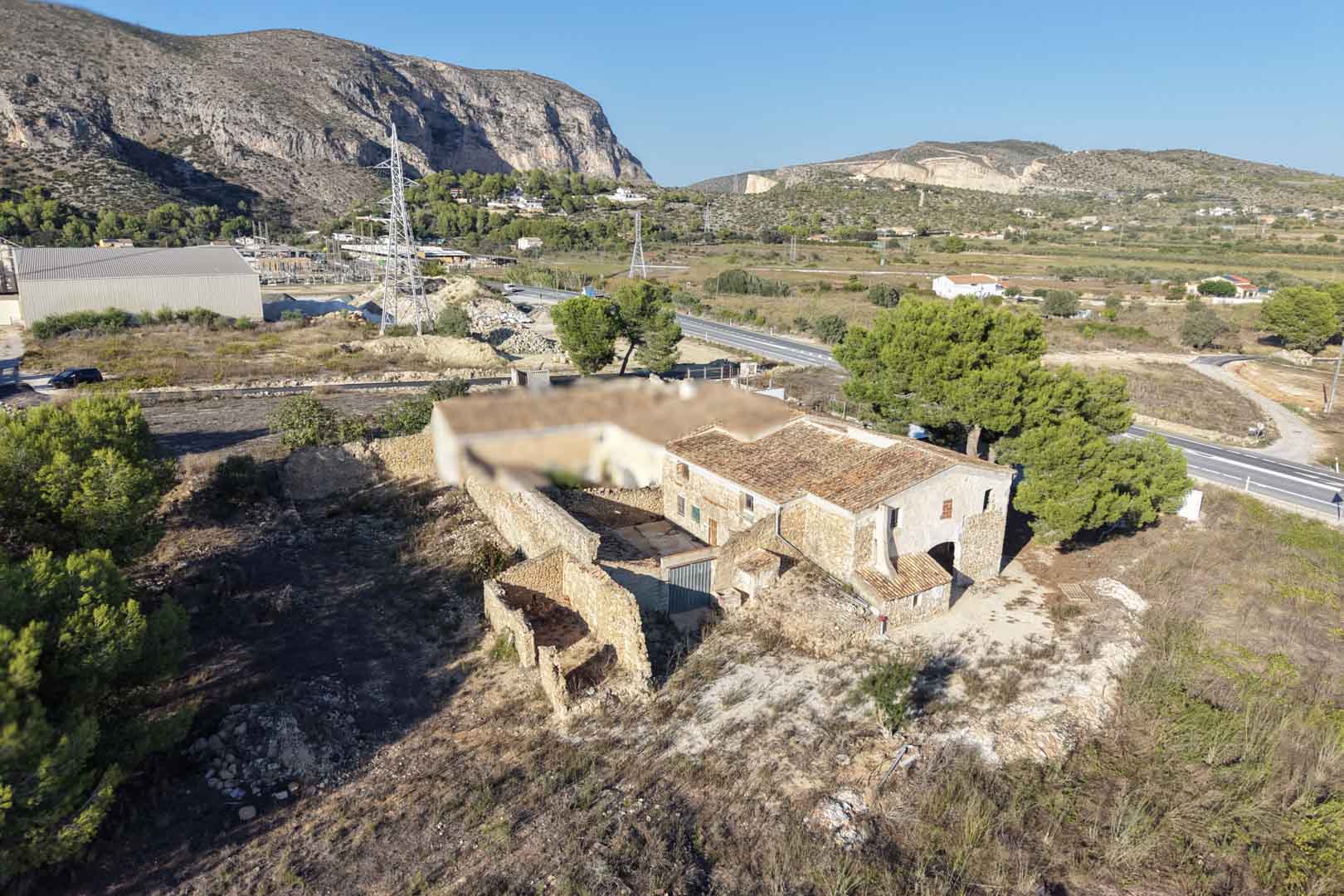 Vista aérea de la casa de piedra tradicional con patio interior y anexos, situada en una extensa parcela rústica con vistas a las montañas de Teulada.