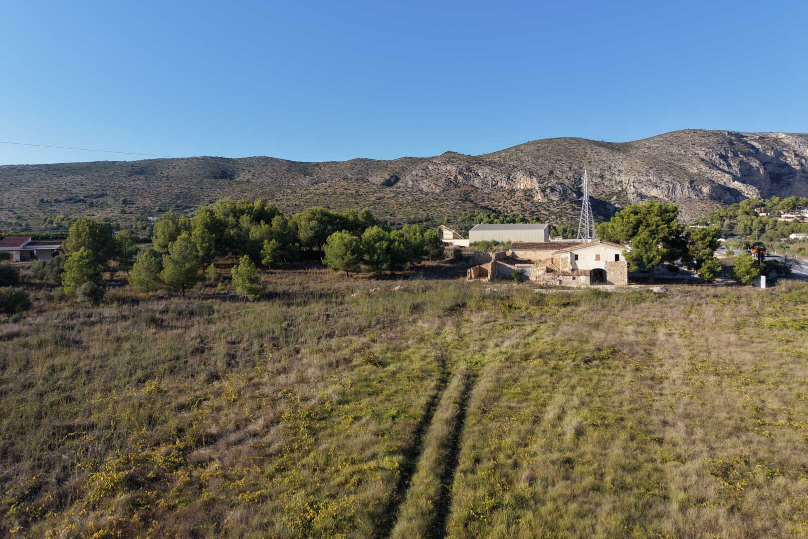 Vista aérea de la casa de piedra tradicional y su extensa parcela llana, rodeada de vegetación mediterránea y con las montañas de Teulada al fondo.