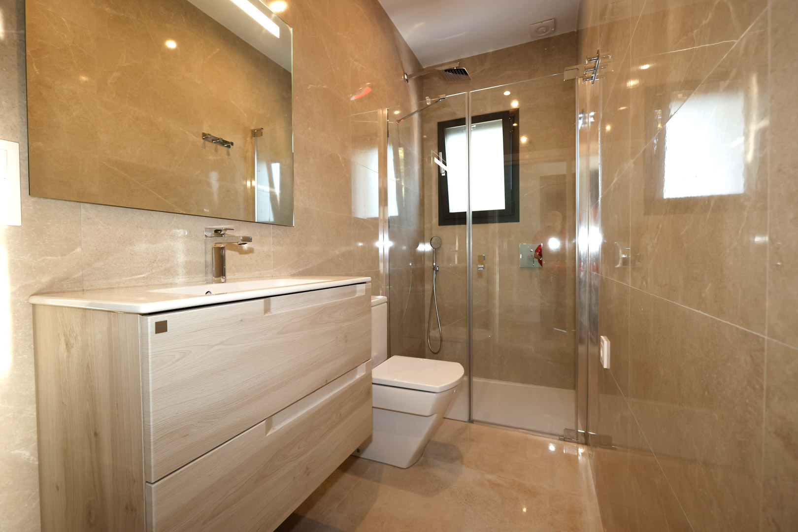 Baño moderno con mueble de lavabo en madera clara, paredes de gres porcelánico efecto mármol y ducha con mampara de cristal y grifería cromada.