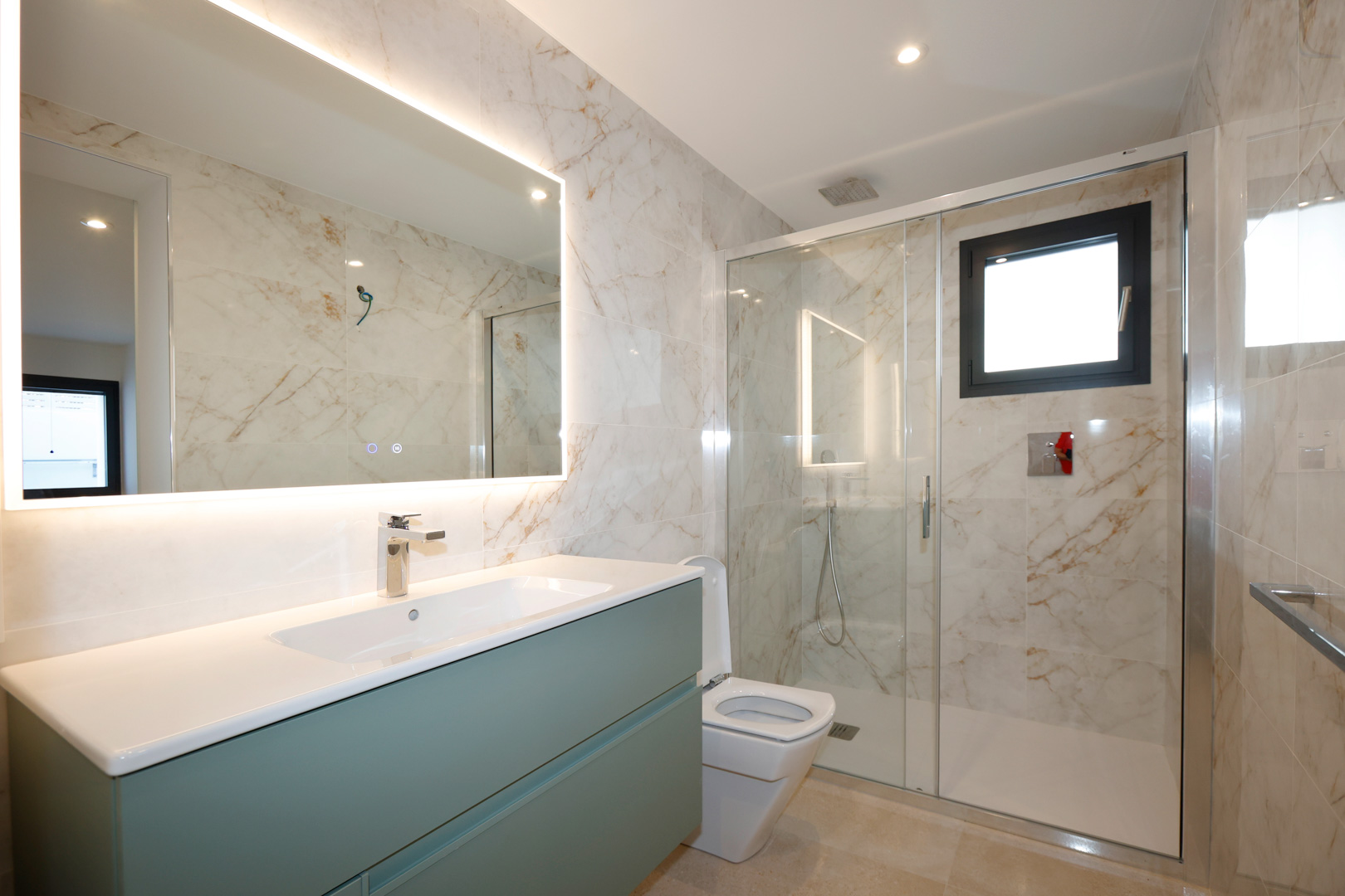 Baño moderno con mueble de lavabo verde, espejo con iluminación LED perimetral y ducha con mampara de cristal y azulejos efecto mármol.