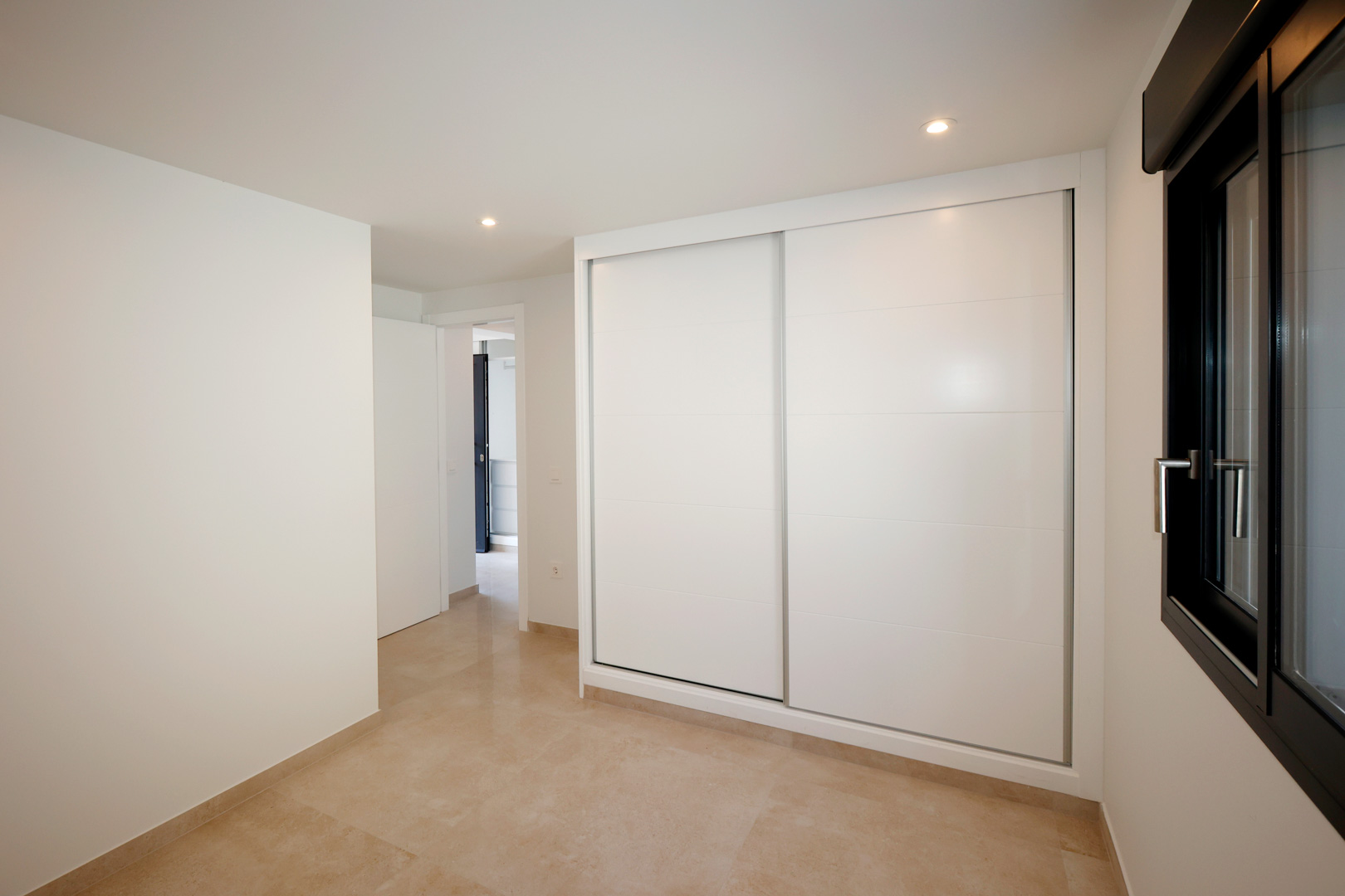 Dormitorio con armario empotrado de puertas correderas blancas, suelo de gres porcelánico e iluminación LED integrada junto a una ventana de aluminio oscuro.