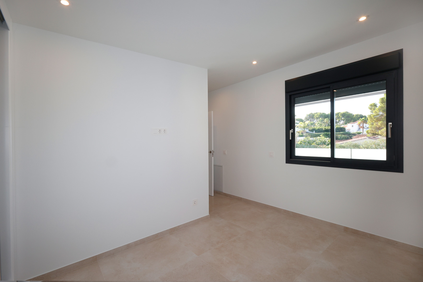 Dormitorio minimalista con paredes blancas, suelo de gres porcelánico y ventana de aluminio negro con persiana eléctrica y vistas al entorno natural.