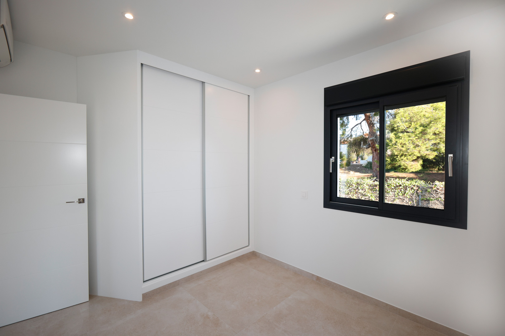 Dormitorio moderno con armario empotrado de puertas correderas blancas, suelo de gres porcelánico y ventana de aluminio negro con vistas al exterior.