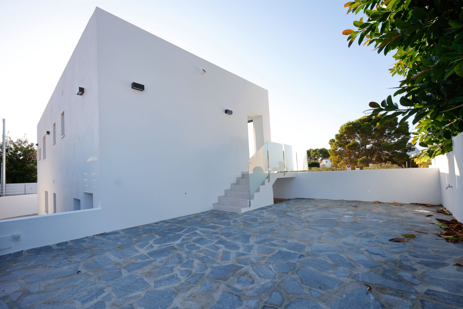 Fachada minimalista blanca con un amplio patio de piedra natural irregular y escalera exterior con barandillas de cristal y acero inoxidable.