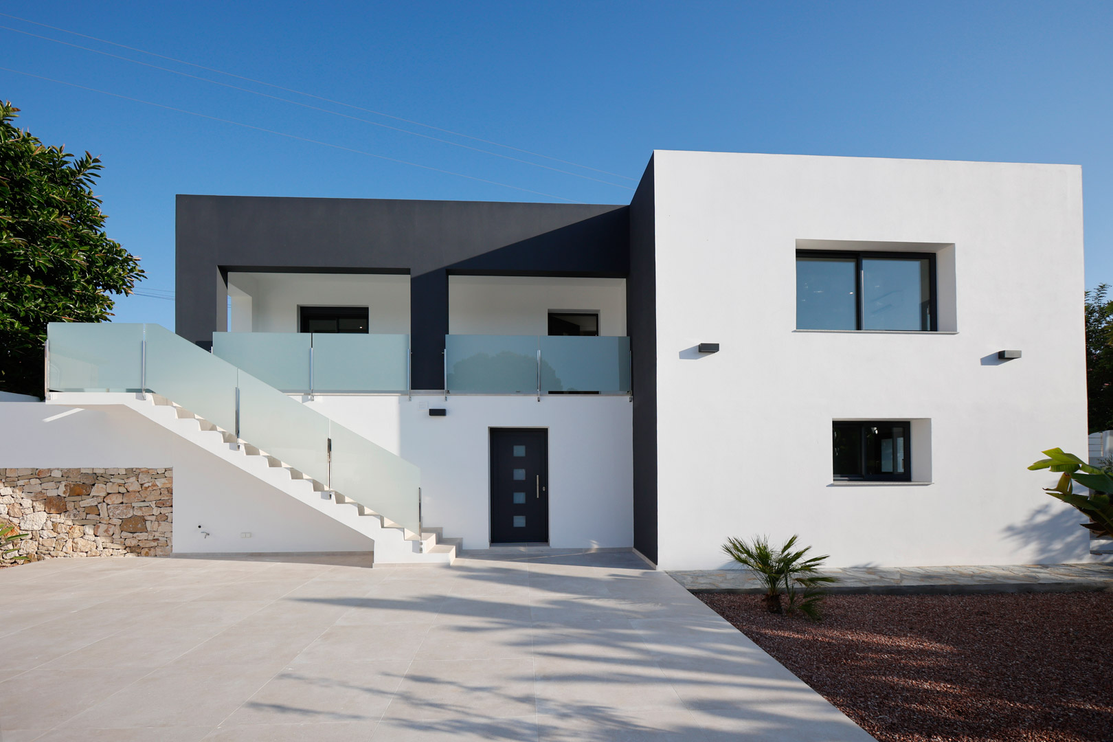 Fachada minimalista en blanco y gris con escalera exterior, barandillas de cristal de seguridad, carpintería Technal y muro perimetral de piedra natural.