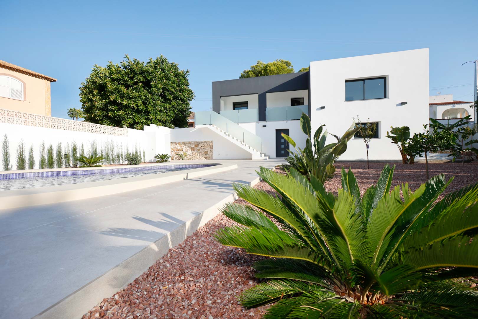 Fachada moderna de dos niveles con piscina, jardín de grava roja y escalera exterior con barandillas de cristal que conecta las viviendas independientes.