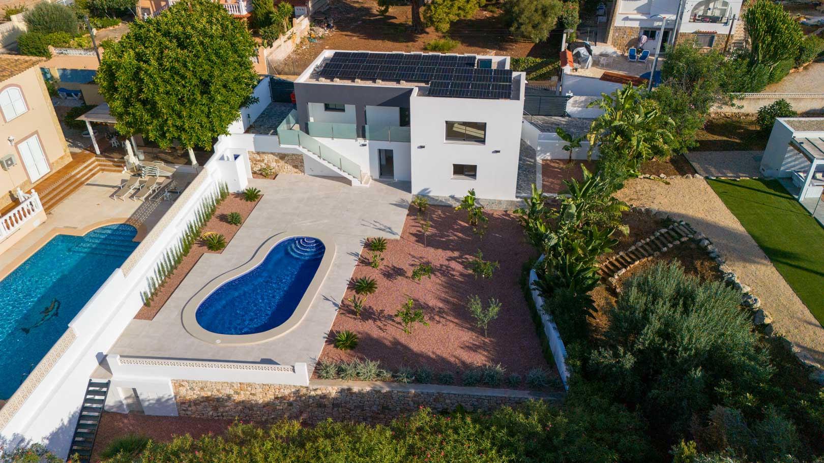 Vista aérea de la villa moderna con paneles solares, piscina de diseño orgánico y jardín de grava marmoleada roja delimitado por muros de piedra natural.