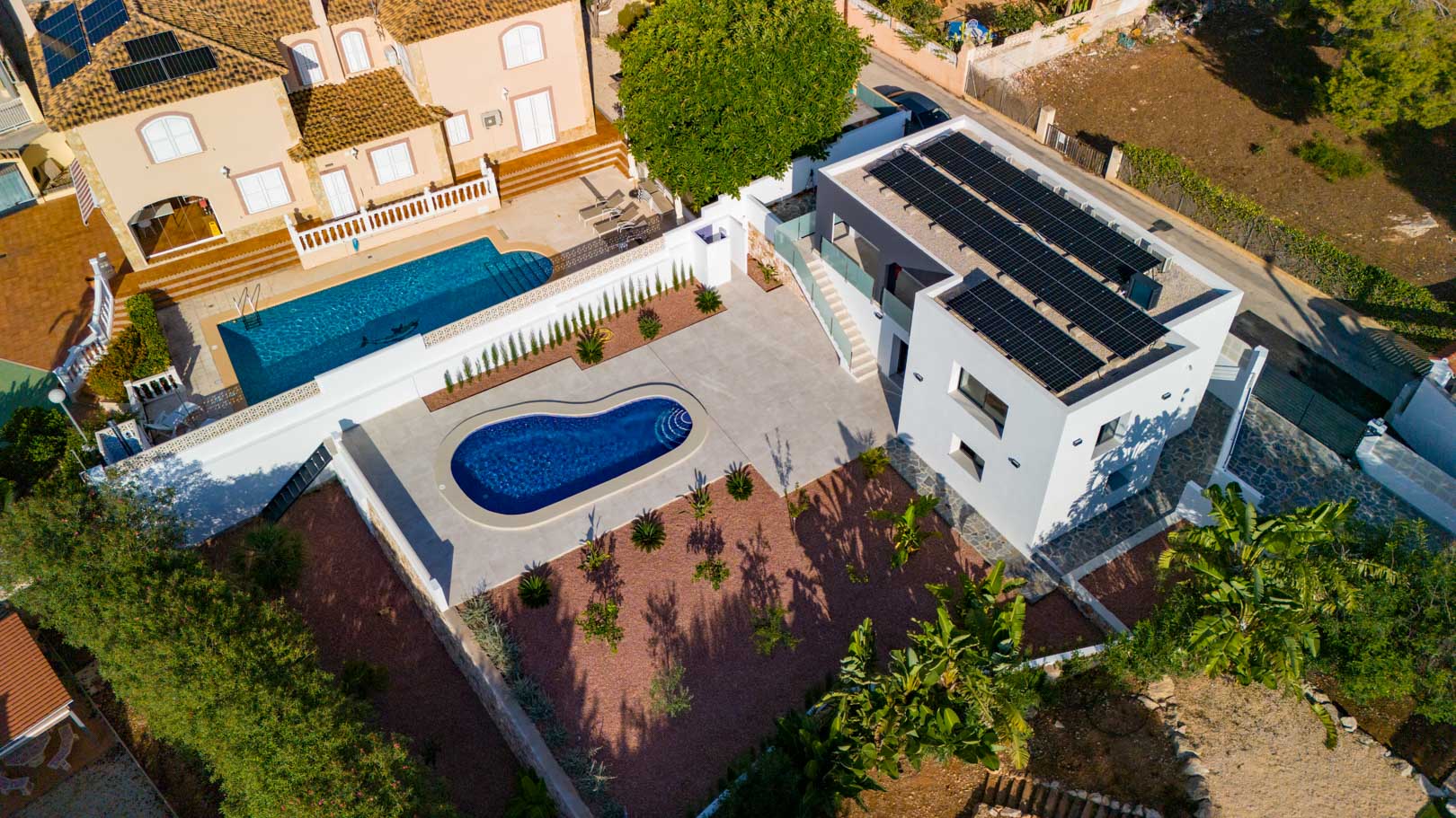 Vista aérea de la villa moderna con paneles solares, piscina de diseño y jardín de grava marmoleada roja. Destacan las barandillas de cristal y los muros de piedra natural.