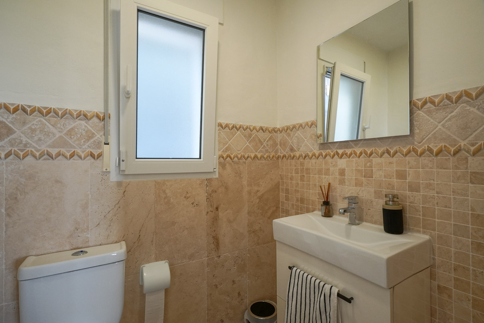 Aseo de cortesía con azulejos en tonos tierra, cenefa decorativa, lavabo moderno y ventana de cristal translúcido que aporta luz natural.