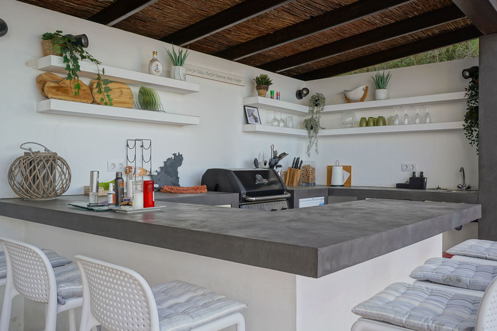 Cocina de verano exterior con encimeras de microcemento gris, barbacoa integrada, estanterías abiertas y techo de cañizo con vigas de madera.