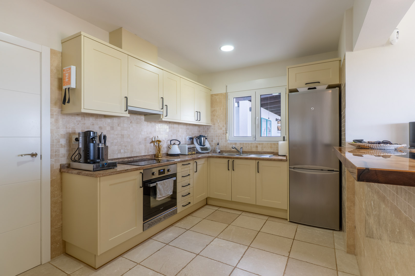 Cocina funcional equipada con mobiliario en tono crema, encimeras de granito y electrodomésticos de acero inoxidable, incluyendo una barra de desayuno en madera maciza.