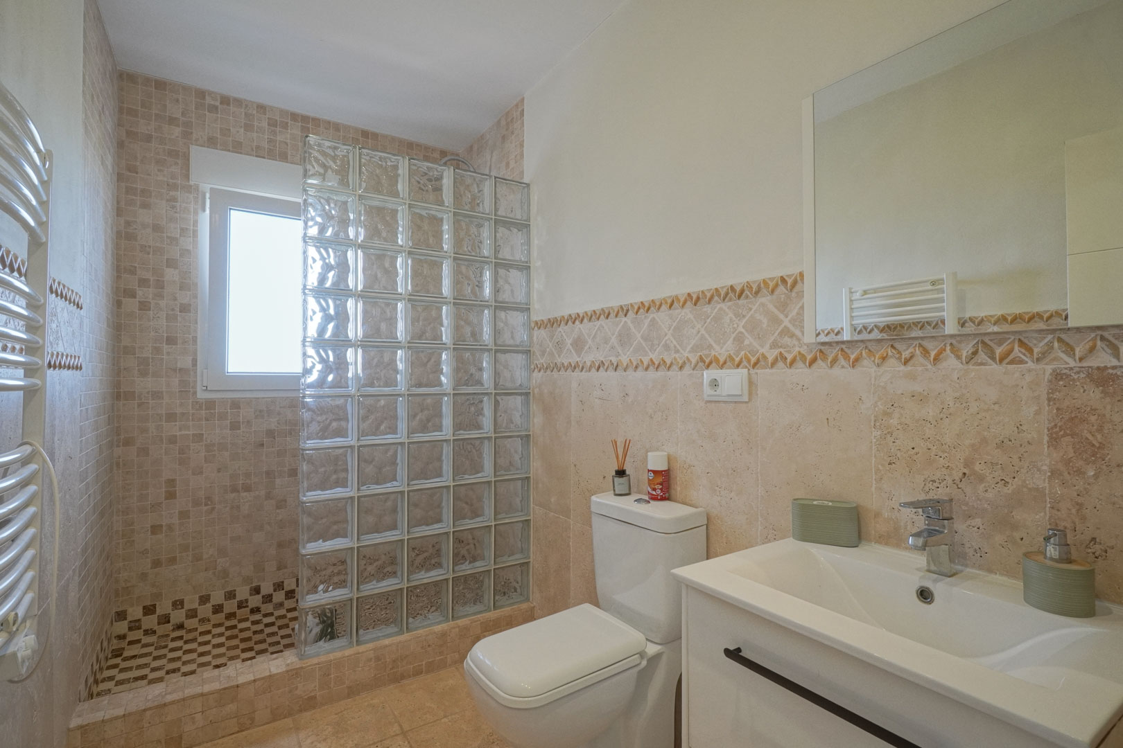 Cuarto de baño con ducha de obra, mampara de bloques de vidrio, azulejos de tonos tierra, lavabo con mueble blanco y radiador toallero.