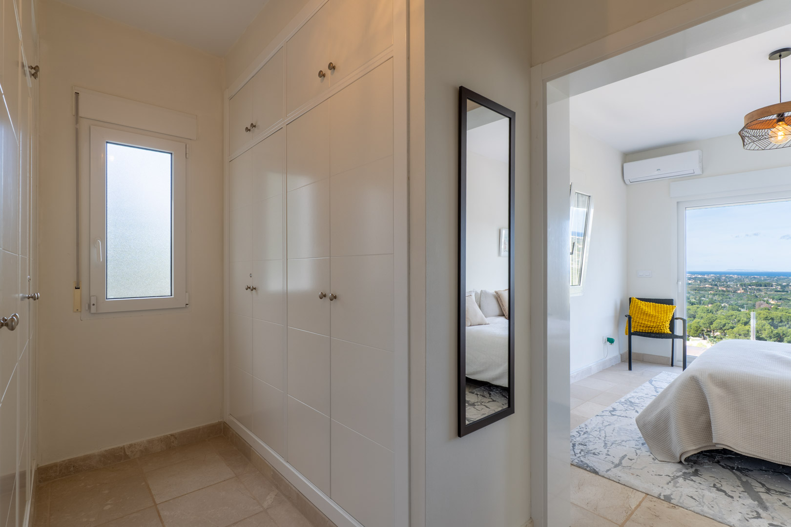 Pasillo con armarios empotrados blancos y espejo de cuerpo entero que conecta con un dormitorio con vistas al mar y aire acondicionado.
