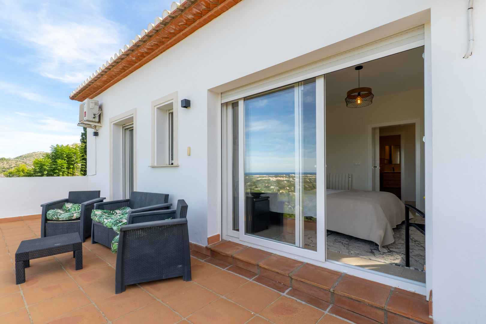 Terraza superior con suelos de terracota y mobiliario de exterior, conectada al dormitorio mediante grandes ventanales con vistas panorámicas al mar Mediterráneo.