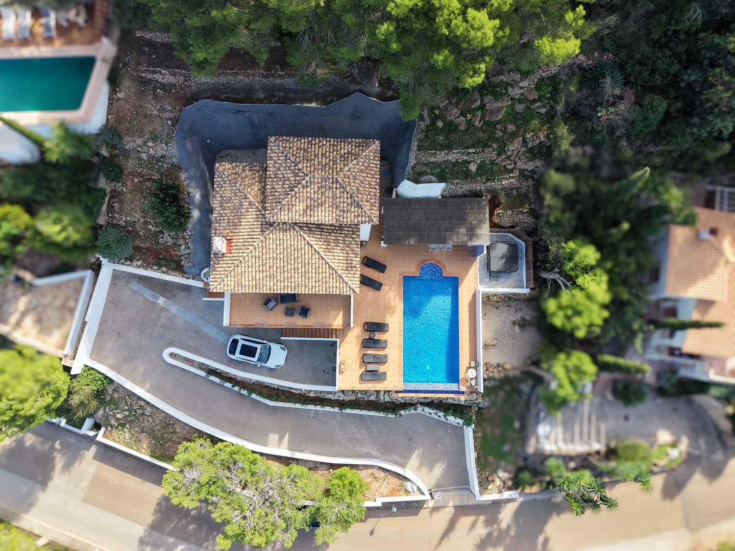 Vista aérea cenital de la villa con tejado de teja mediterránea, piscina rectangular, terraza con tumbonas, jacuzzi exterior y zona de aparcamiento privada rodeada de pinos.