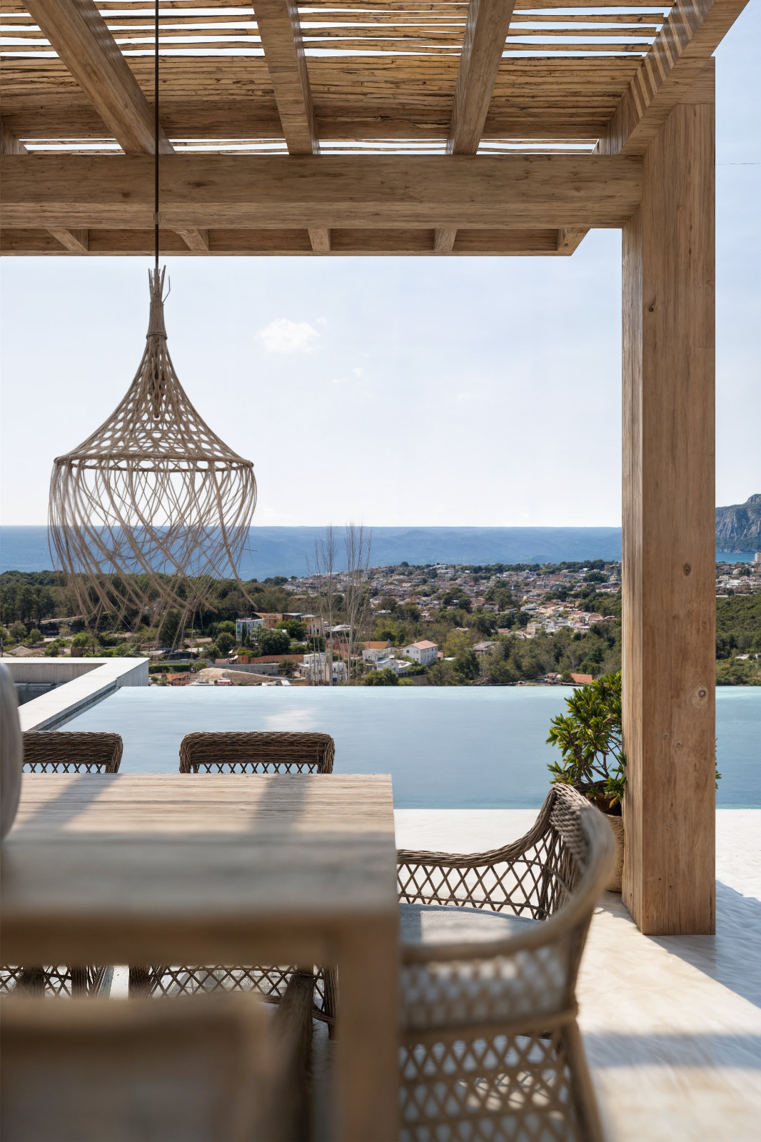 Terraza exterior con pérgola de madera y cañizo, comedor con sillas de mimbre y vistas panorámicas al mar sobre la piscina infinita.
