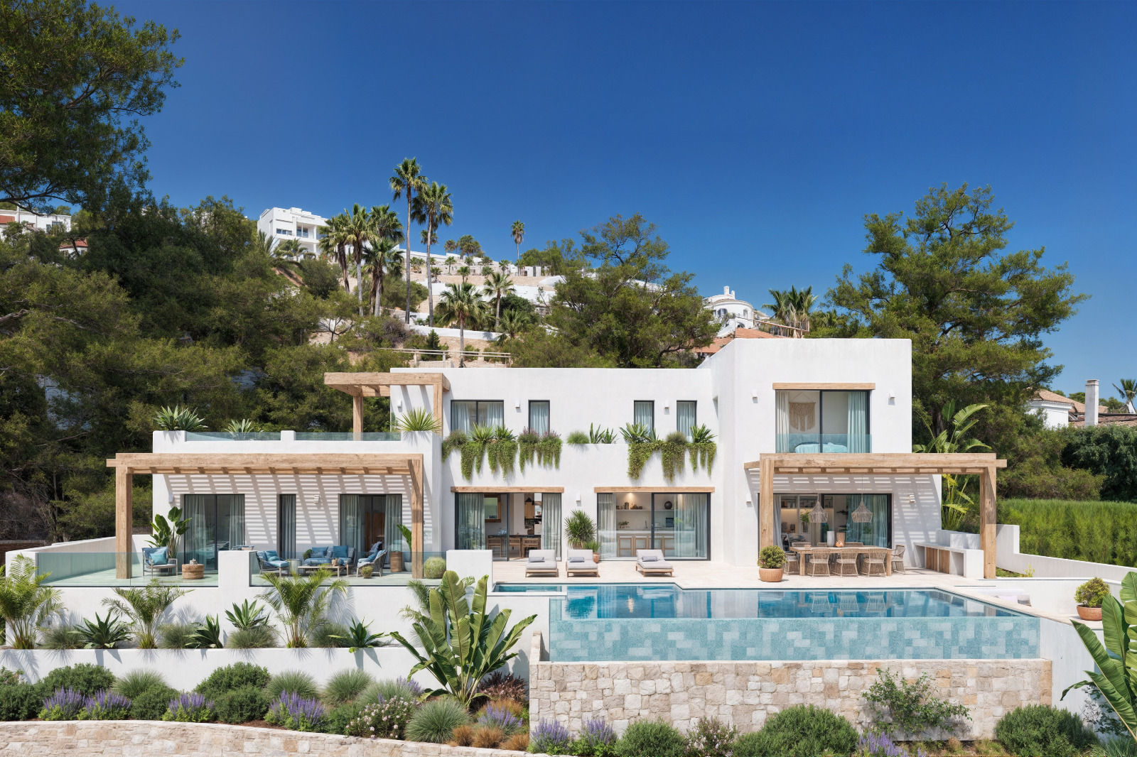 Vista exterior de la villa con fachadas blancas, pérgolas de madera, piscina infinita y jardín mediterráneo con muros de piedra natural.