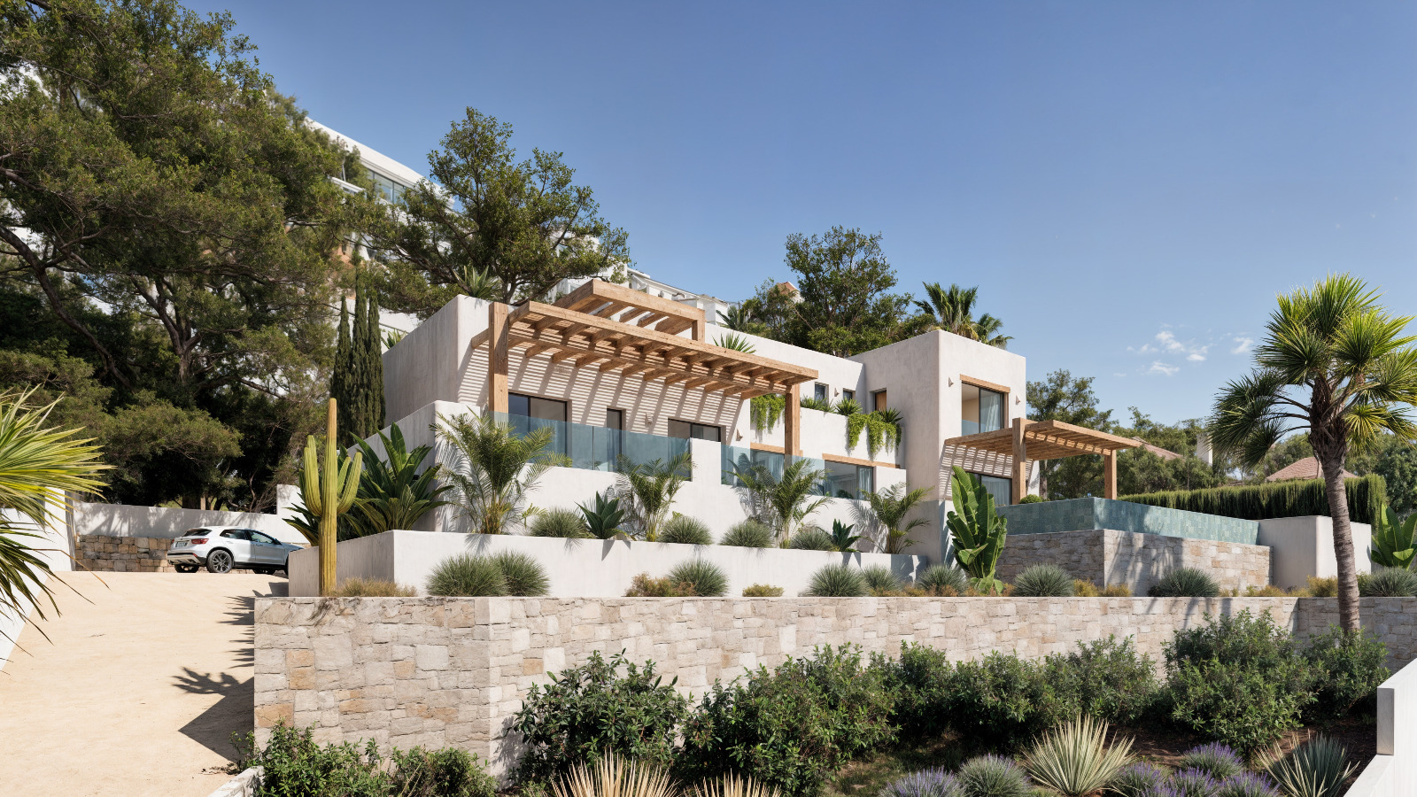 Vista exterior de la villa de estilo ibicenco con fachadas blancas, pérgolas de madera y muros de piedra natural integrados en un jardín mediterráneo con palmeras.
