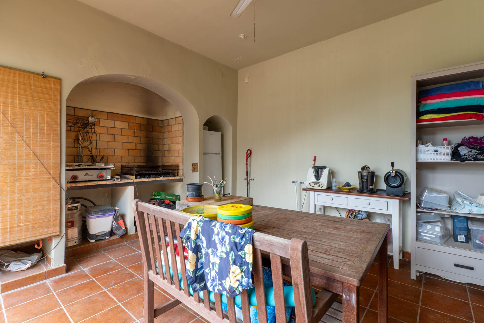 Cocina exterior con barbacoa de obra integrada en arco de ladrillo, mesa de madera rústica y suelos de terracota tradicionales.
