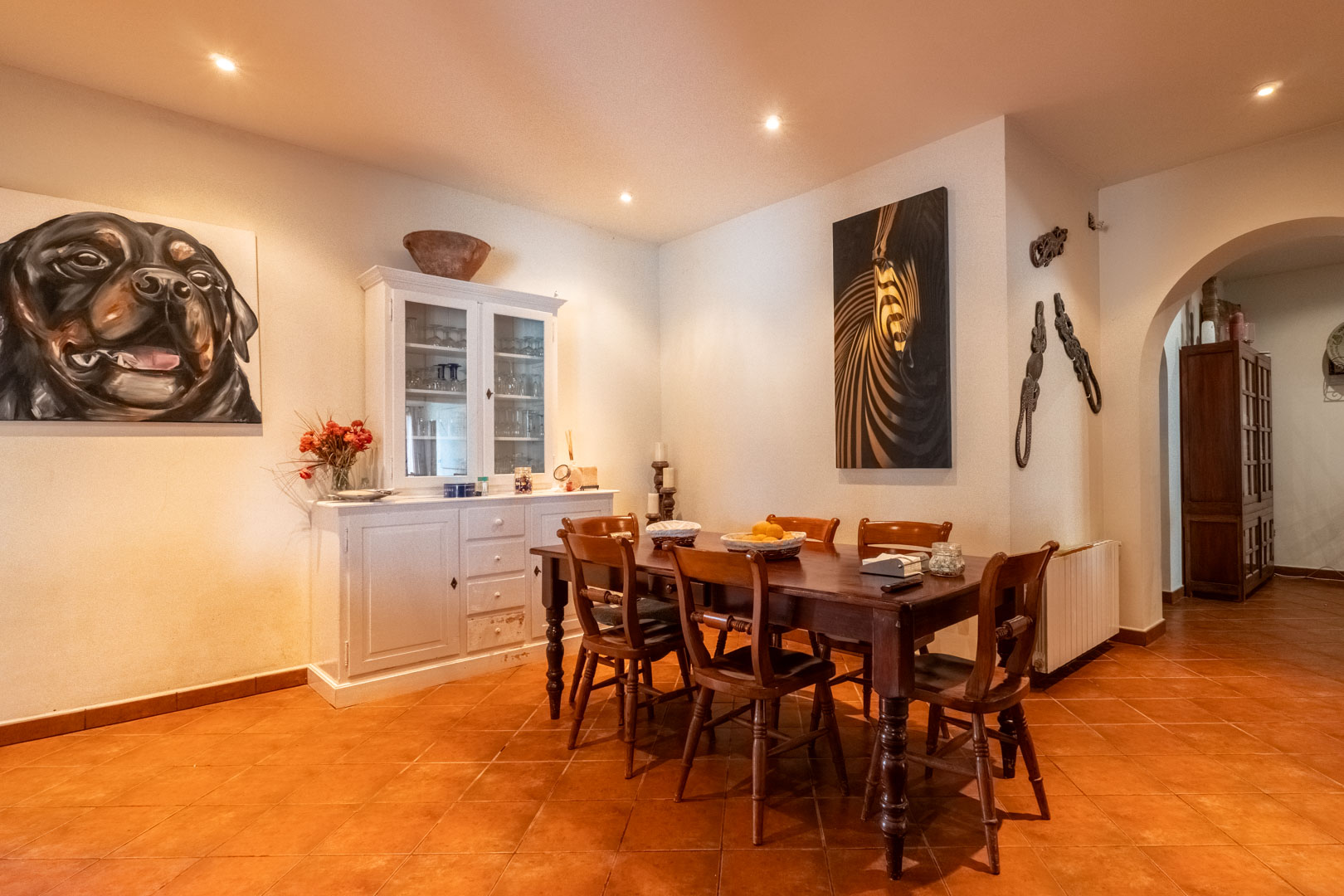 Comedor con mesa de madera oscura, sillas a juego y suelo de terracota. Incluye un aparador blanco clásico, iluminación empotrada y acceso mediante arco a otras estancias.