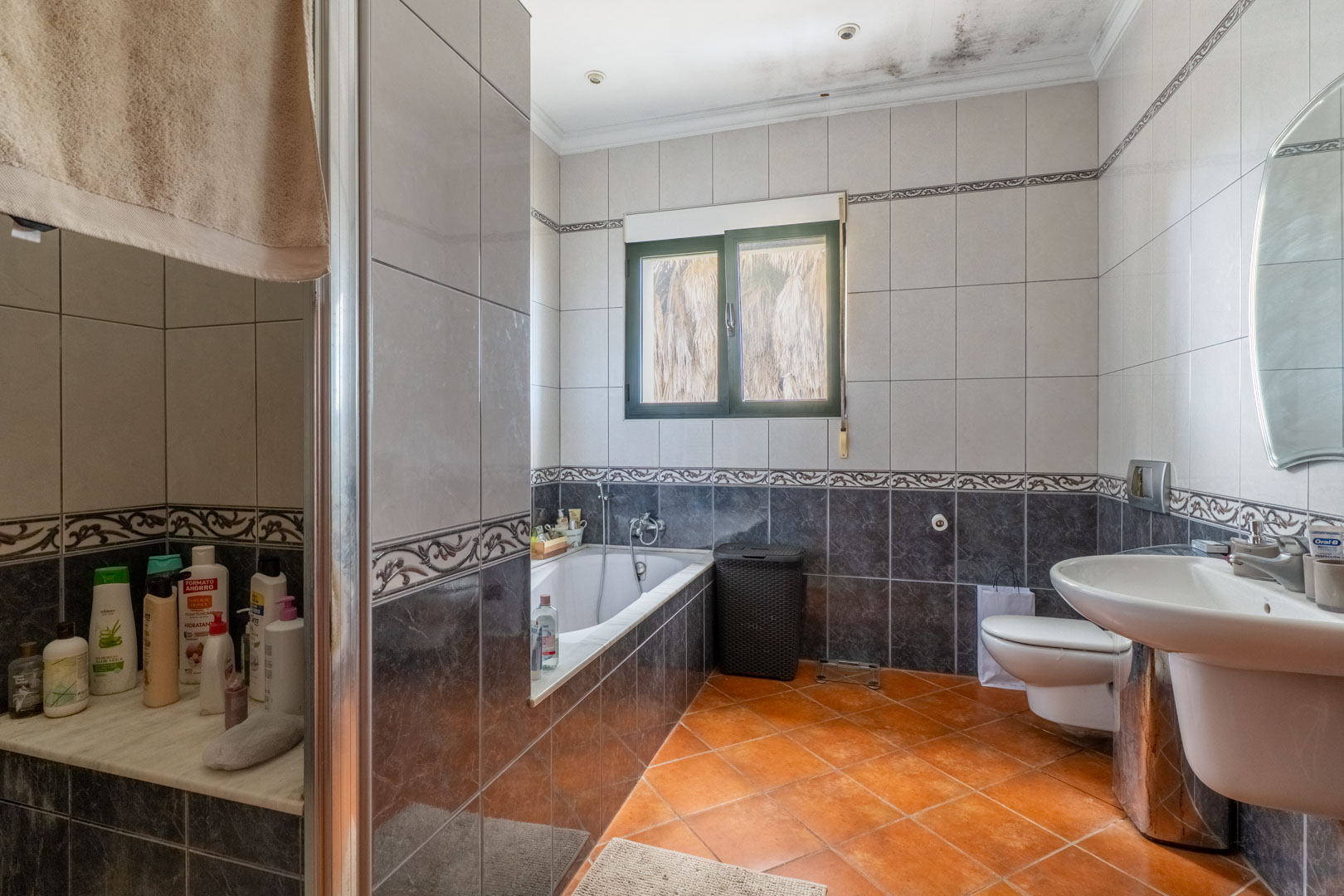 Cuarto de baño con azulejos bitono, bañera encastrada, suelo de terracota y ventana. Incluye sanitario suspendido, lavabo de porcelana y espejo ovalado.