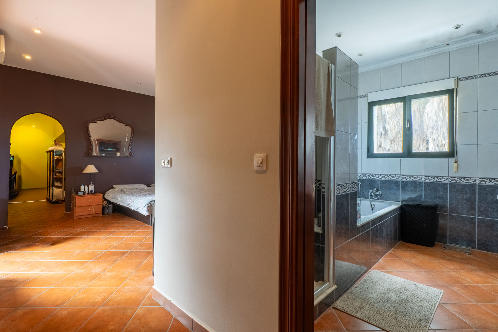 Dormitorio con suelos de barro cocido y pared de acento oscura, conectado a un baño en suite alicatado en tonos grises con bañera integrada y luz natural.