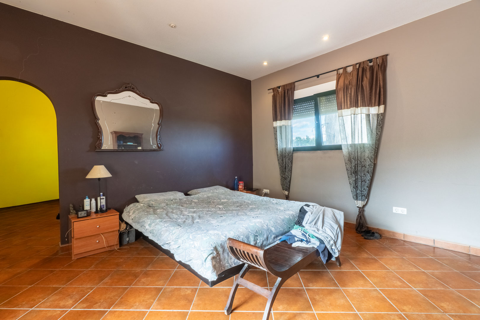 Dormitorio con suelos de terracota, pared de acento chocolate y un arco que comunica con otra estancia. Incluye iluminación empotrada y ventana con persianas.