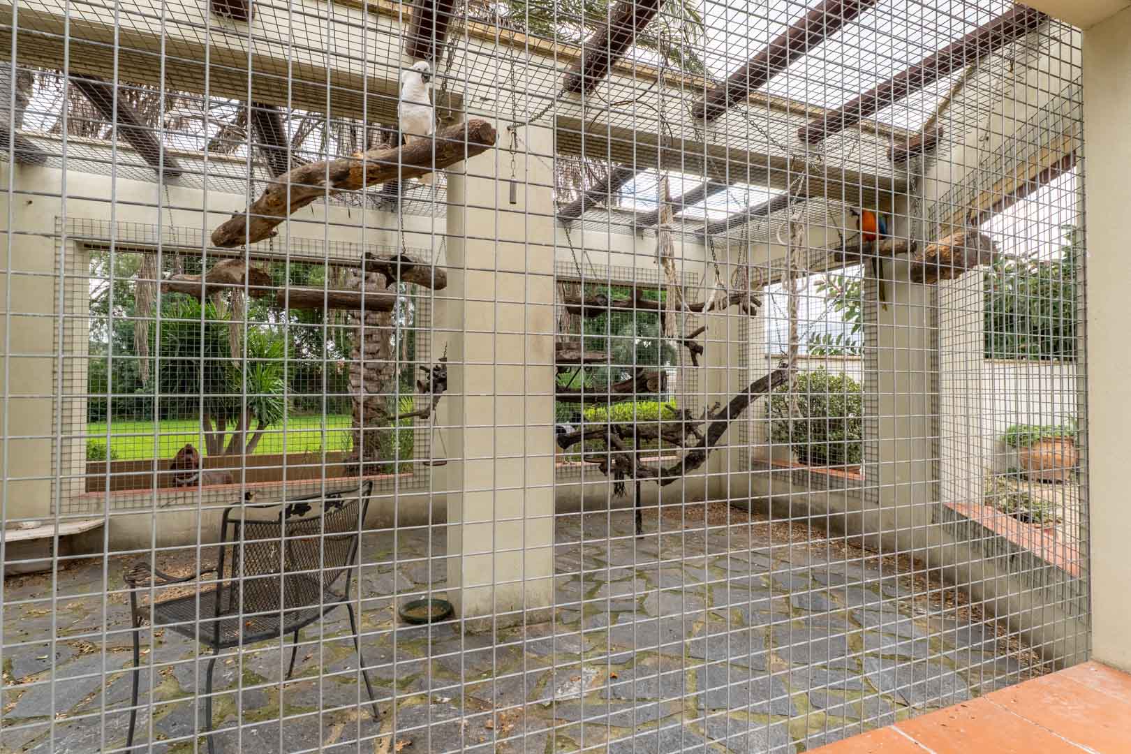 Gran aviario exterior con estructura de malla metálica, suelo de pizarra y perchas de madera natural, integrado en los jardines de la finca.