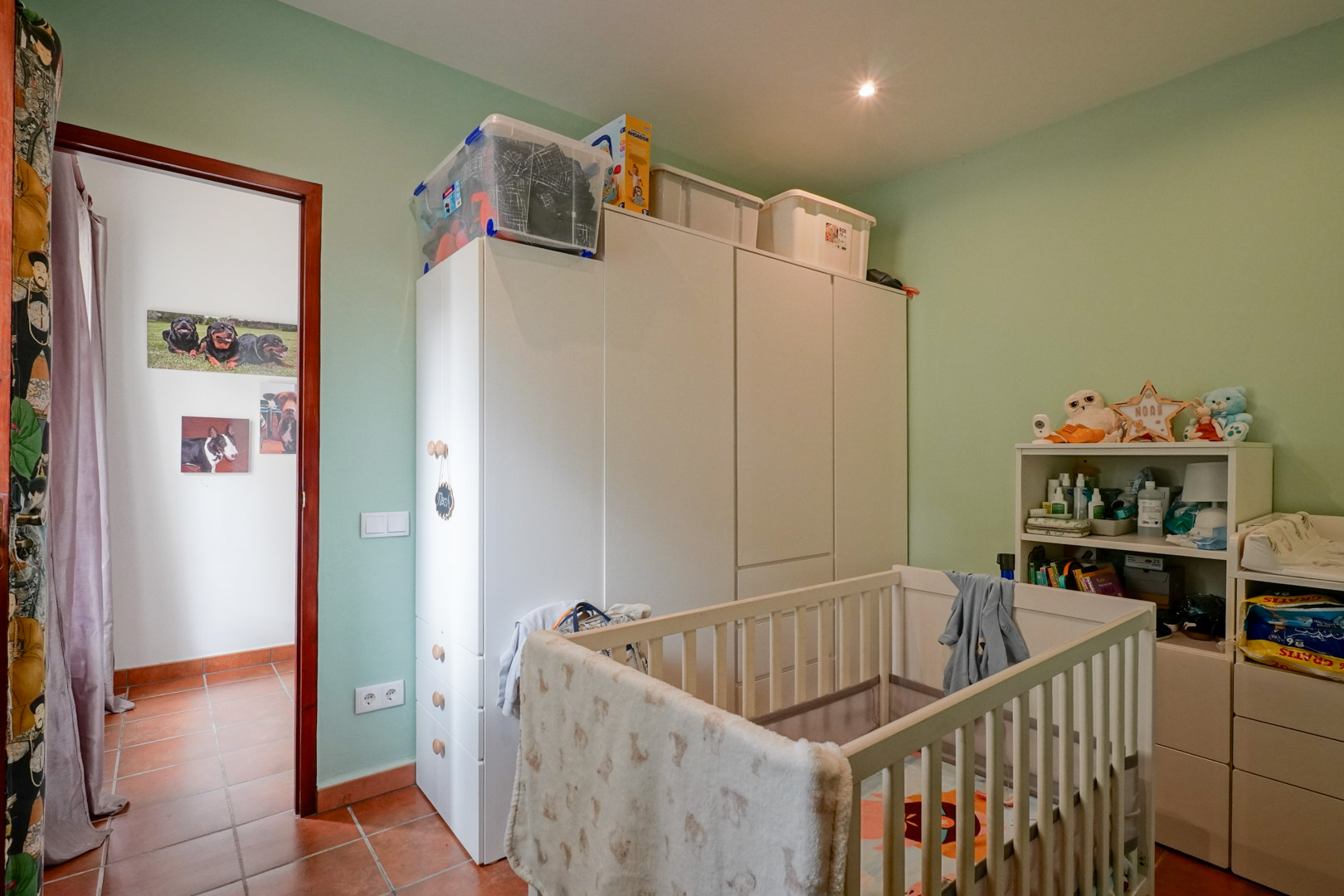 Habitación infantil con cuna de madera blanca, armarios de almacenamiento y paredes en tono verde pastel. El suelo es de gres cerámico y cuenta con iluminación empotrada.