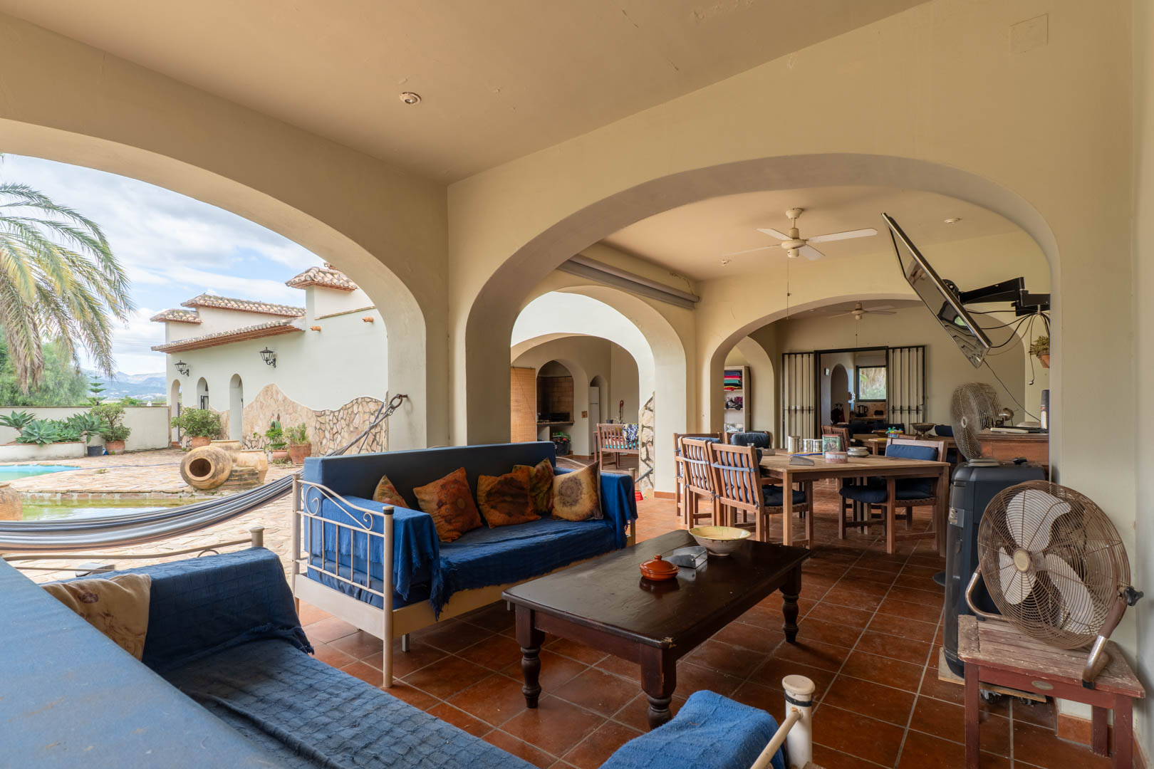 Porche cubierto con arcos mediterráneos, suelo de terracota y zona de estar. Ofrece vistas directas a la piscina, el jardín de palmeras y una zona de comedor exterior.