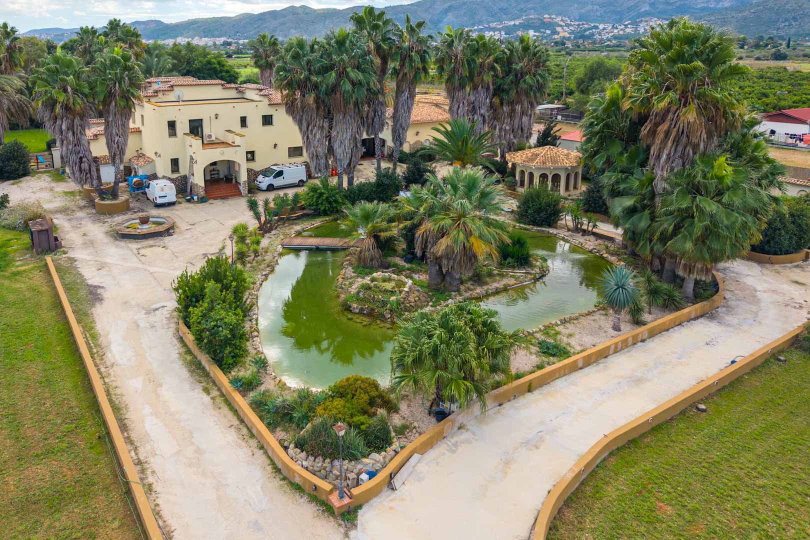 Vista aérea de la entrada principal con estanque ornamental, puentes de madera y palmeras maduras frente a la fachada de estilo mediterráneo.