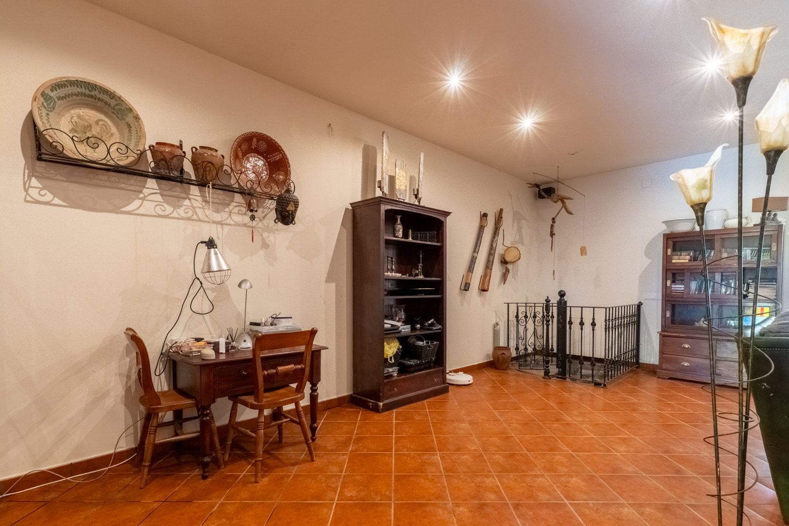 Zona de despacho con escritorio de madera, suelos de terracota y acceso a la planta inferior mediante escalera con barandilla de hierro forjado.